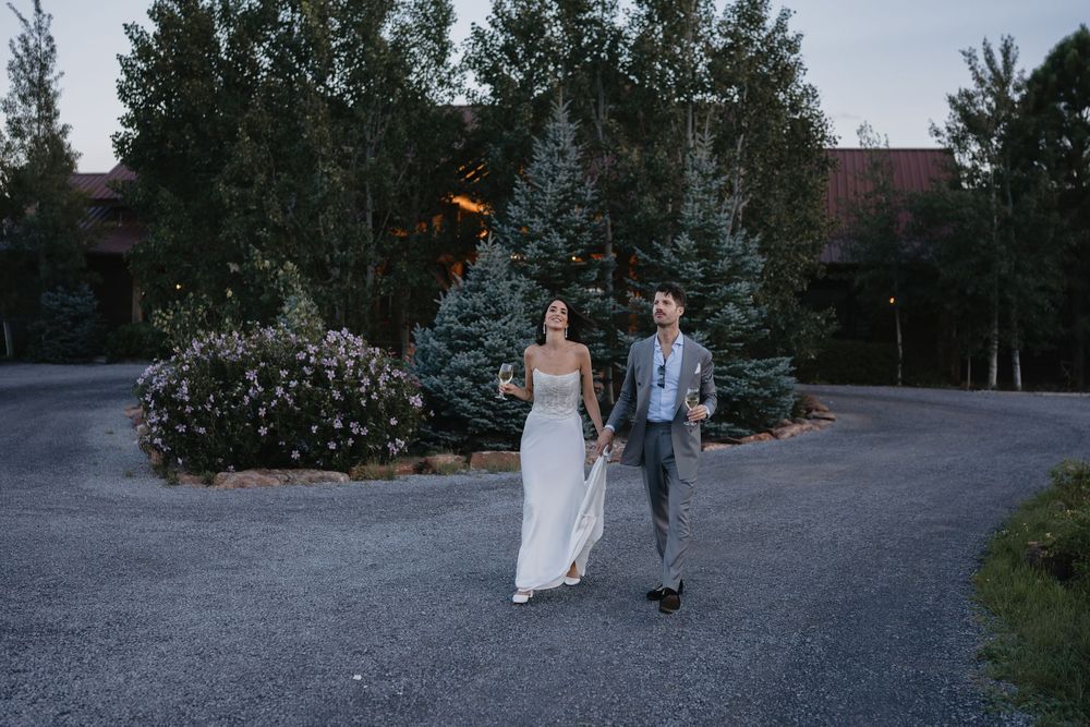 Bride and groom walk hand in hand along a driveway surrounded by lush evergreen trees at dusk.