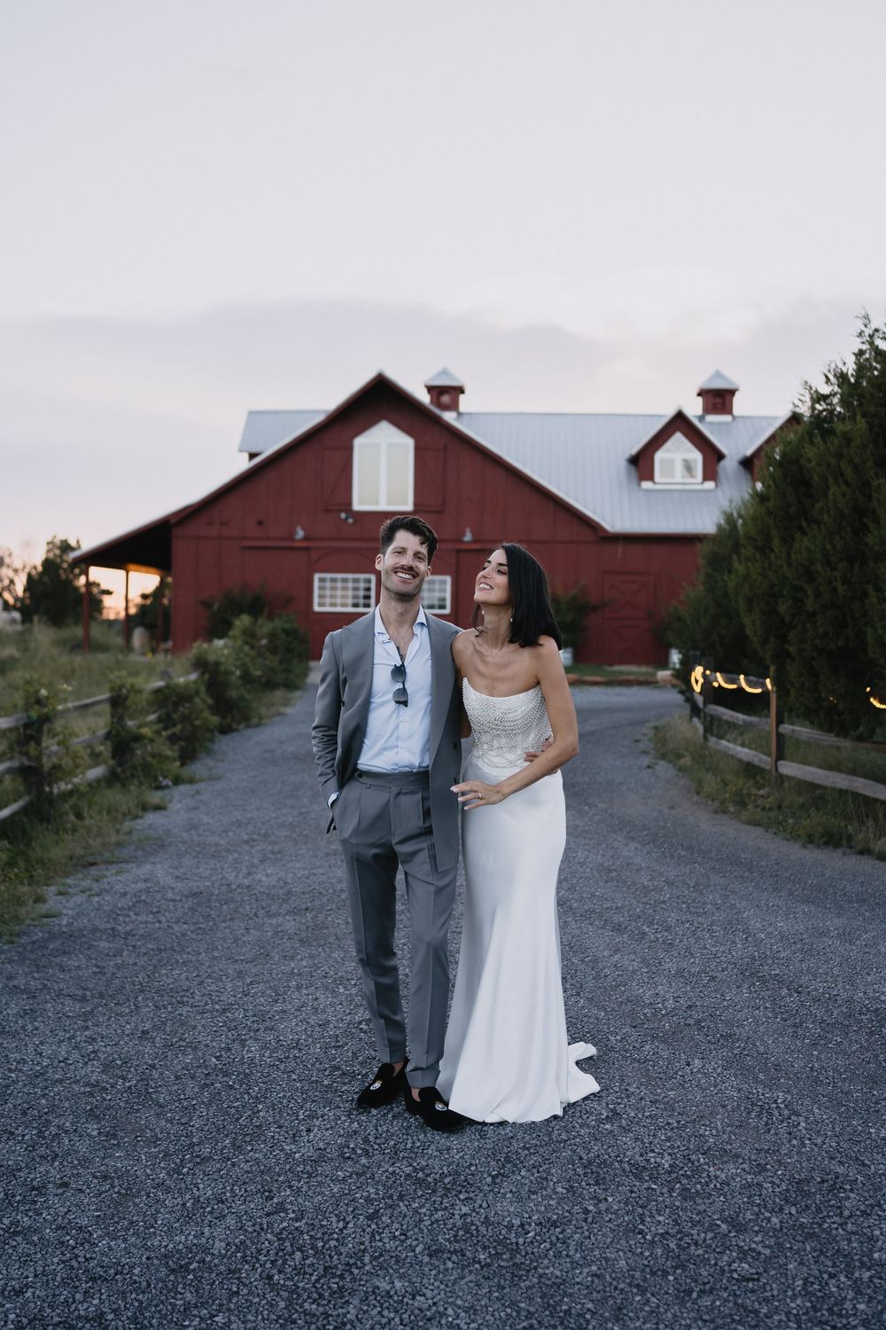 Bride and groom laughing together on gravel path before red barn wedding venue at dusk.