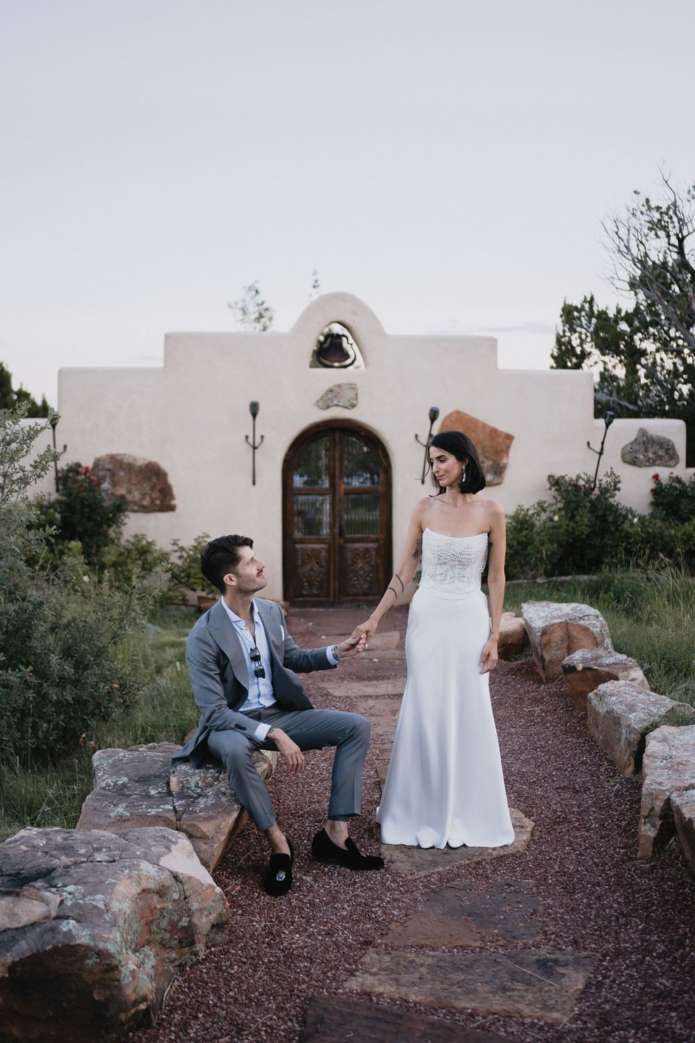 Bride and groom hold hands outside a rustic adobe chapel at dusk, surrounded by natural stone landscaping.