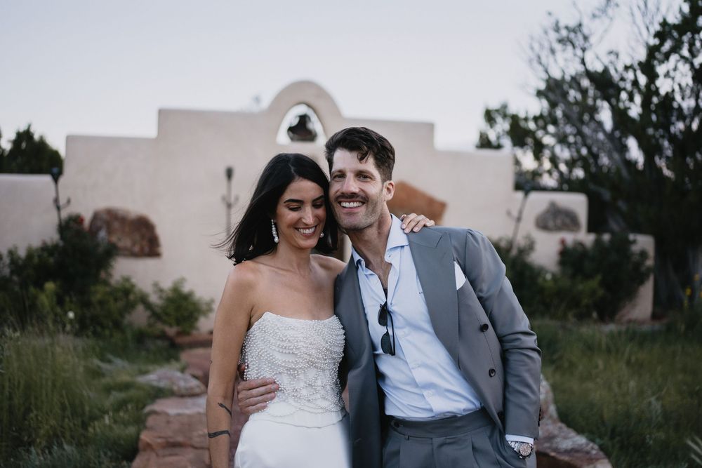 A smiling bride and groom pose together outdoors near a white adobe chapel at dusk.