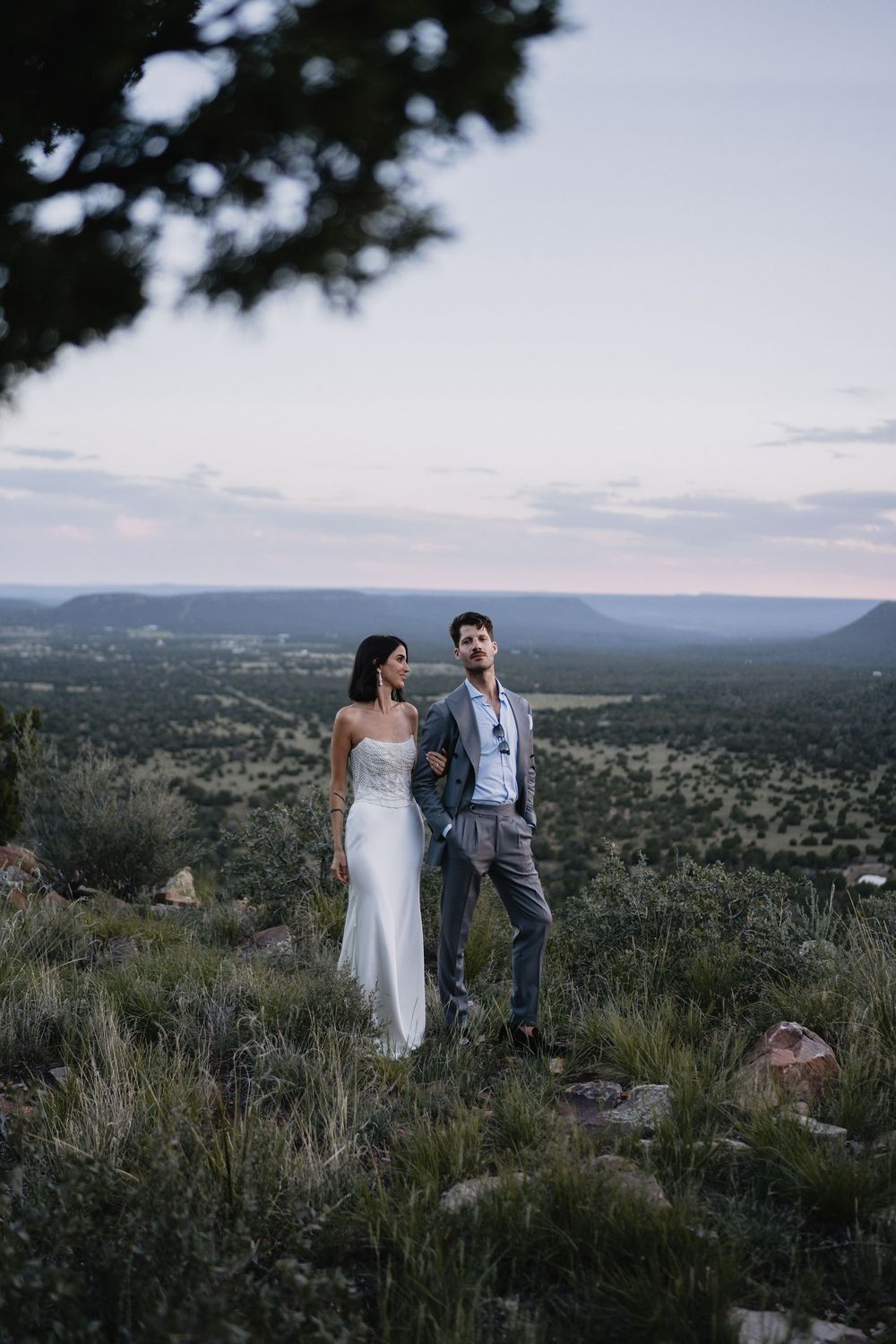Wedding couple stands on a hilltop at dusk overlooking a vast green valley landscape in formal attire.