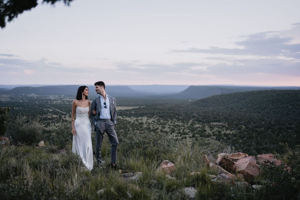Bride and groom stand on a scenic hilltop at dusk, overlooking a vast green valley with dramatic cliffs.