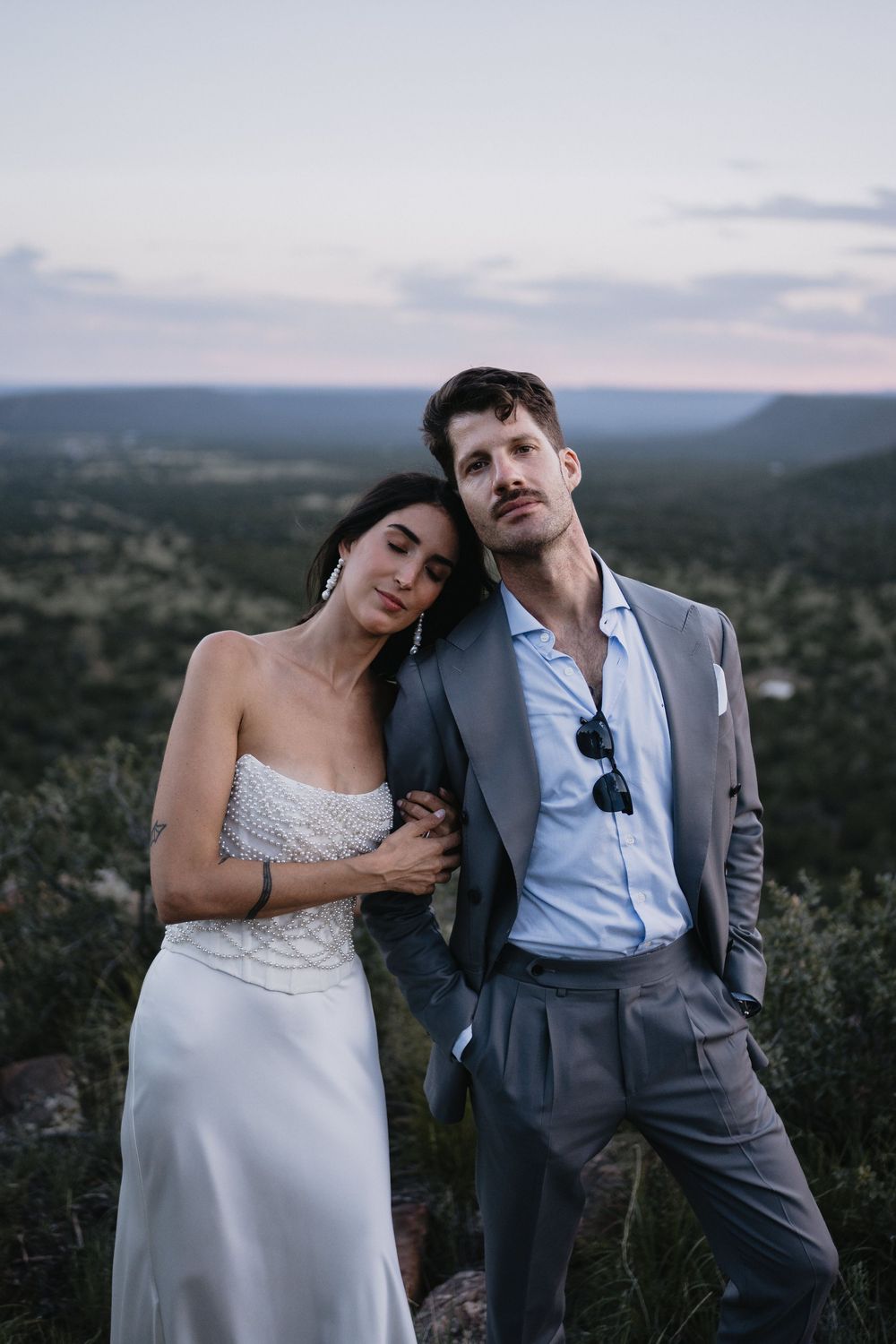Bride and groom pose elegantly at dusk on a scenic overlook, she in white gown, he in gray suit.