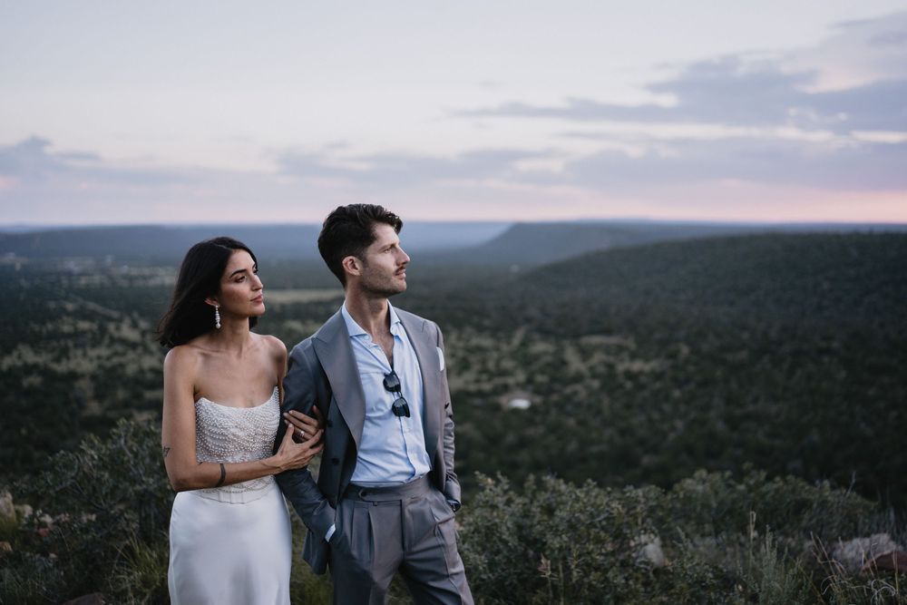 Couple in elegant wedding attire standing on a scenic overlook at dusk with lush forest and pastel sky.
