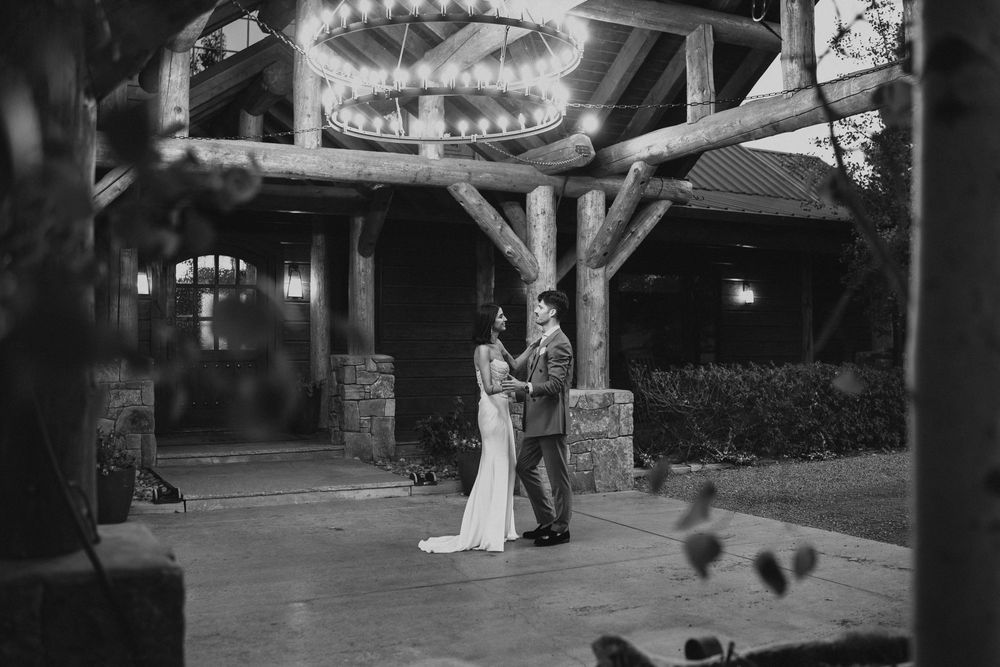 Black and white wedding photo of couple standing under rustic wooden arch with chandelier at elegant outdoor venue.