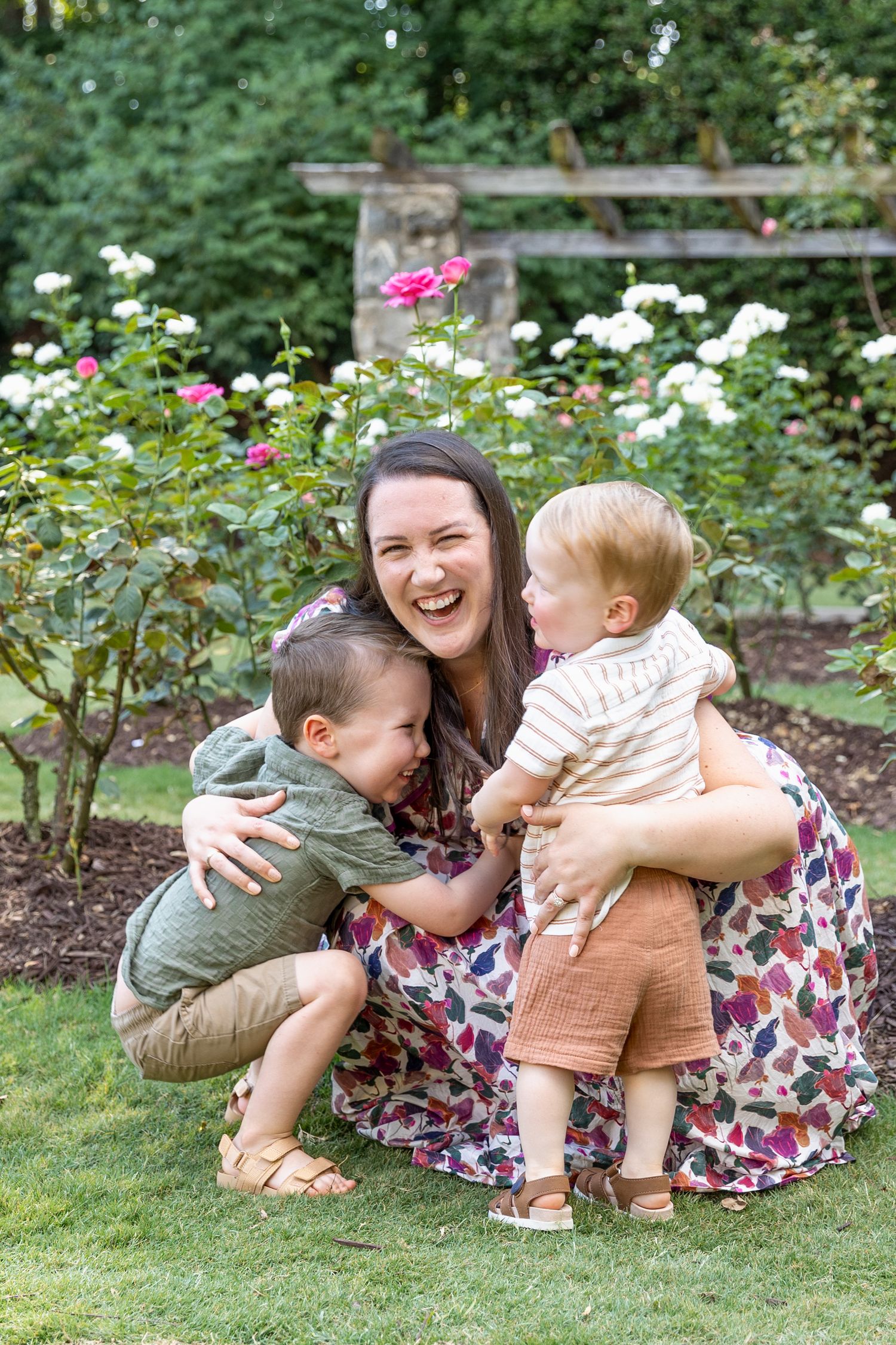 A laughing mother crouches in a garden hugging two young boys near blooming roses.