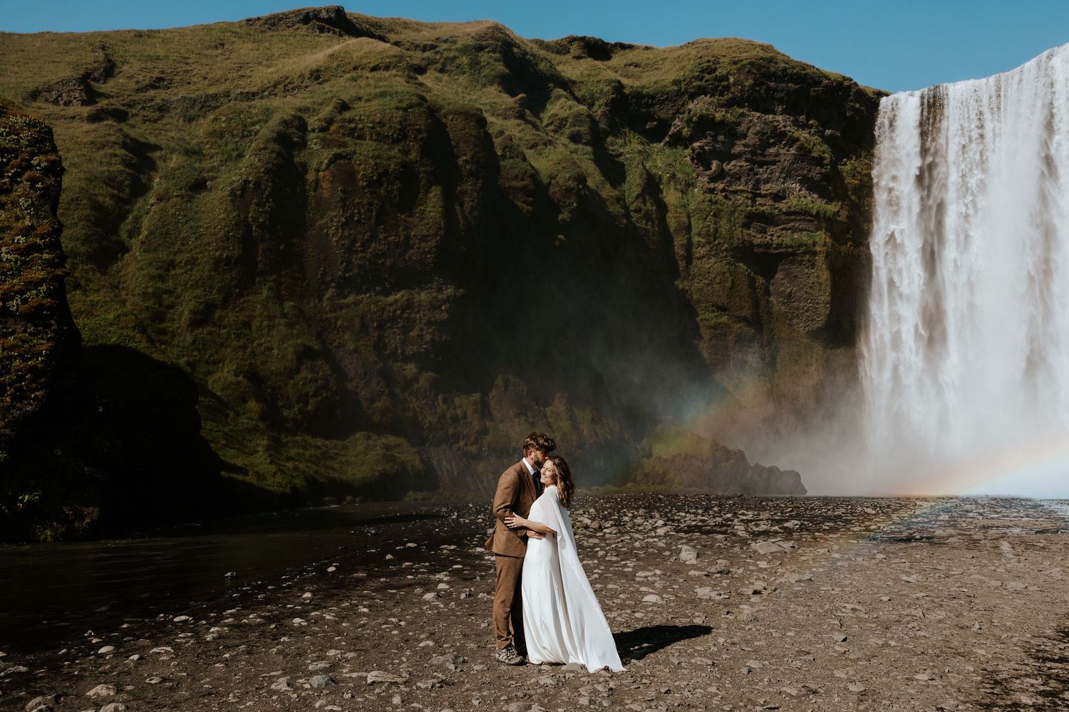 eloping couple snuggling up at Skógafoss in Iceland in front of the waterfall and rainbows