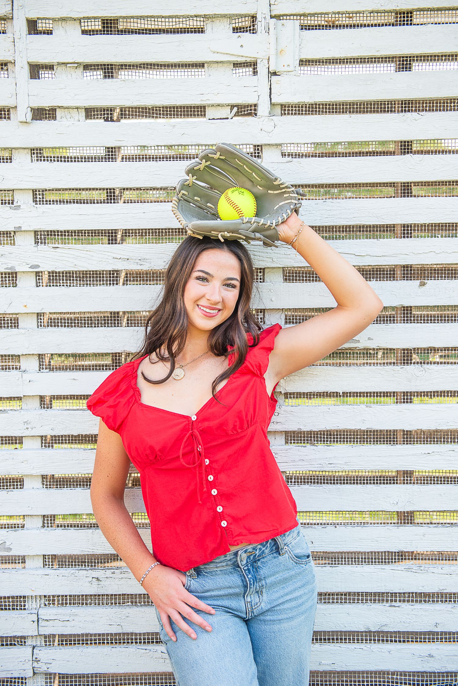 Young woman in red top and jeans holds softball glove overhead, smiling against white lattice fence.