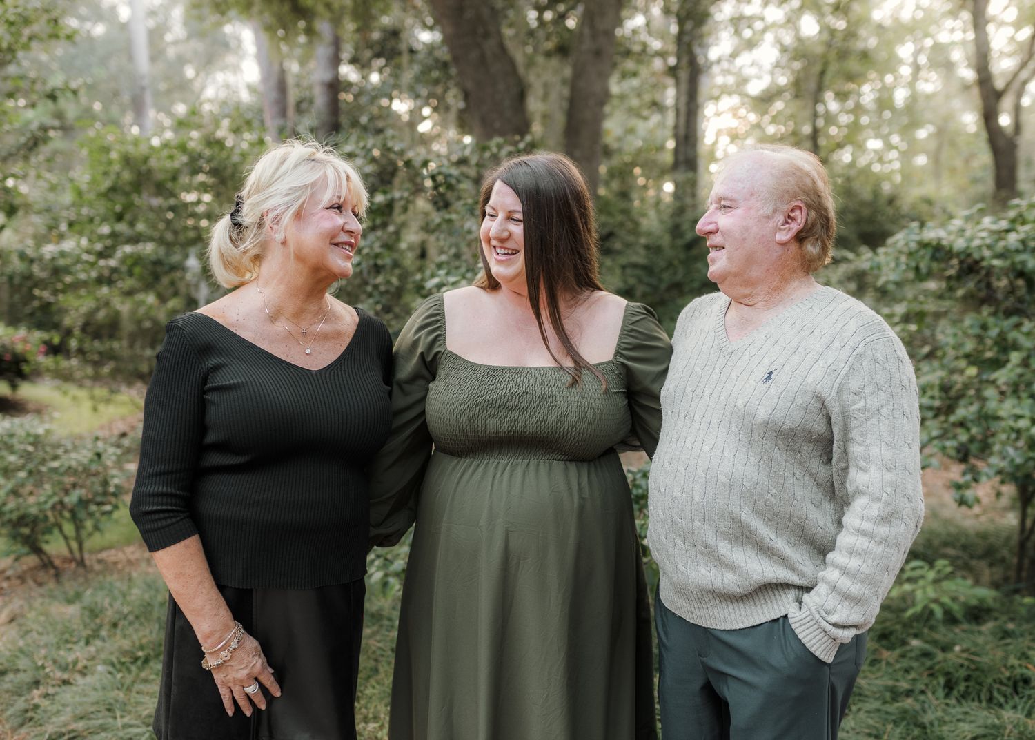 A pregnant woman in a green dress smiles with an older couple outdoors among lush green trees.