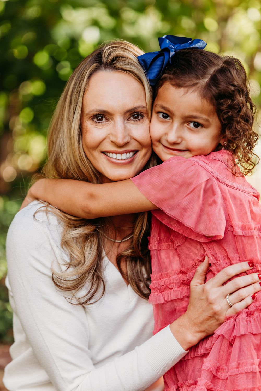 A woman in white hugs a young girl in a pink dress with a blue bow, smiling outdoors among green trees.