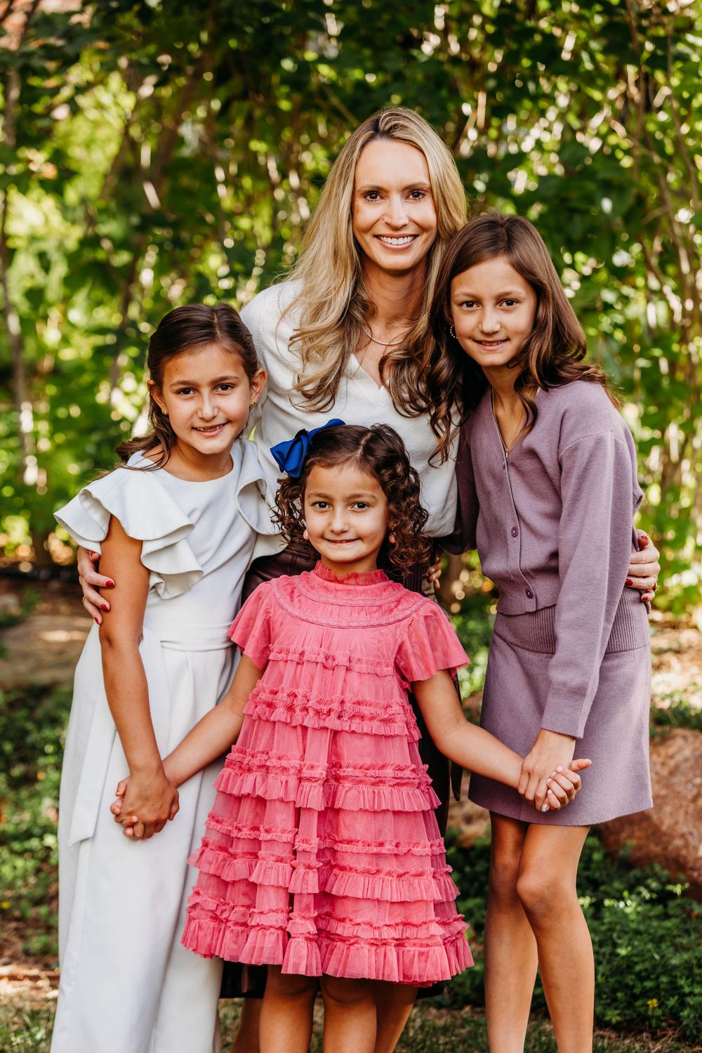 A woman with blonde hair poses outdoors with three young girls in elegant dresses, smiling together in a lush green setting.