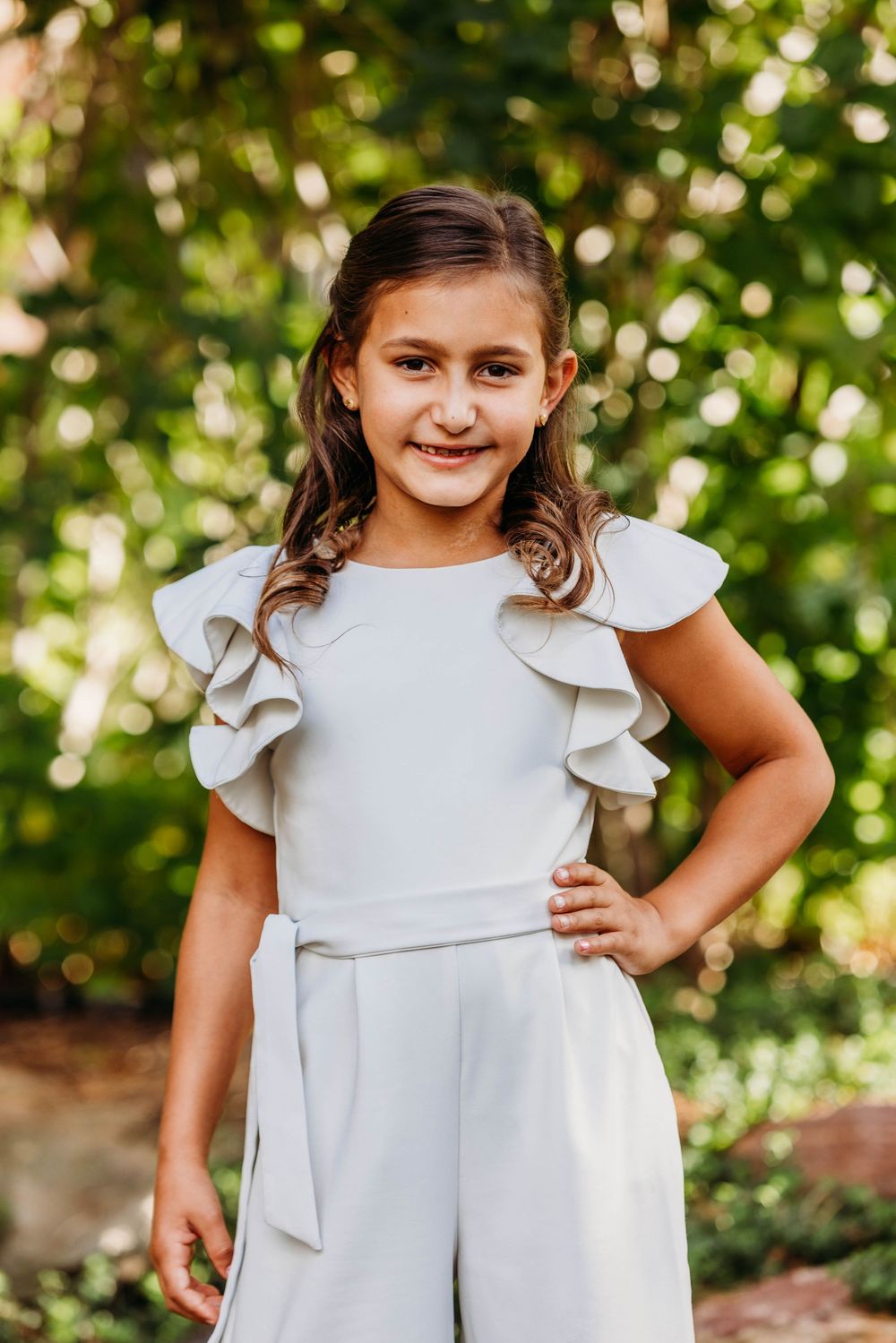 Young girl in white ruffled jumpsuit posing confidently outdoors with hand on hip, surrounded by green foliage.
