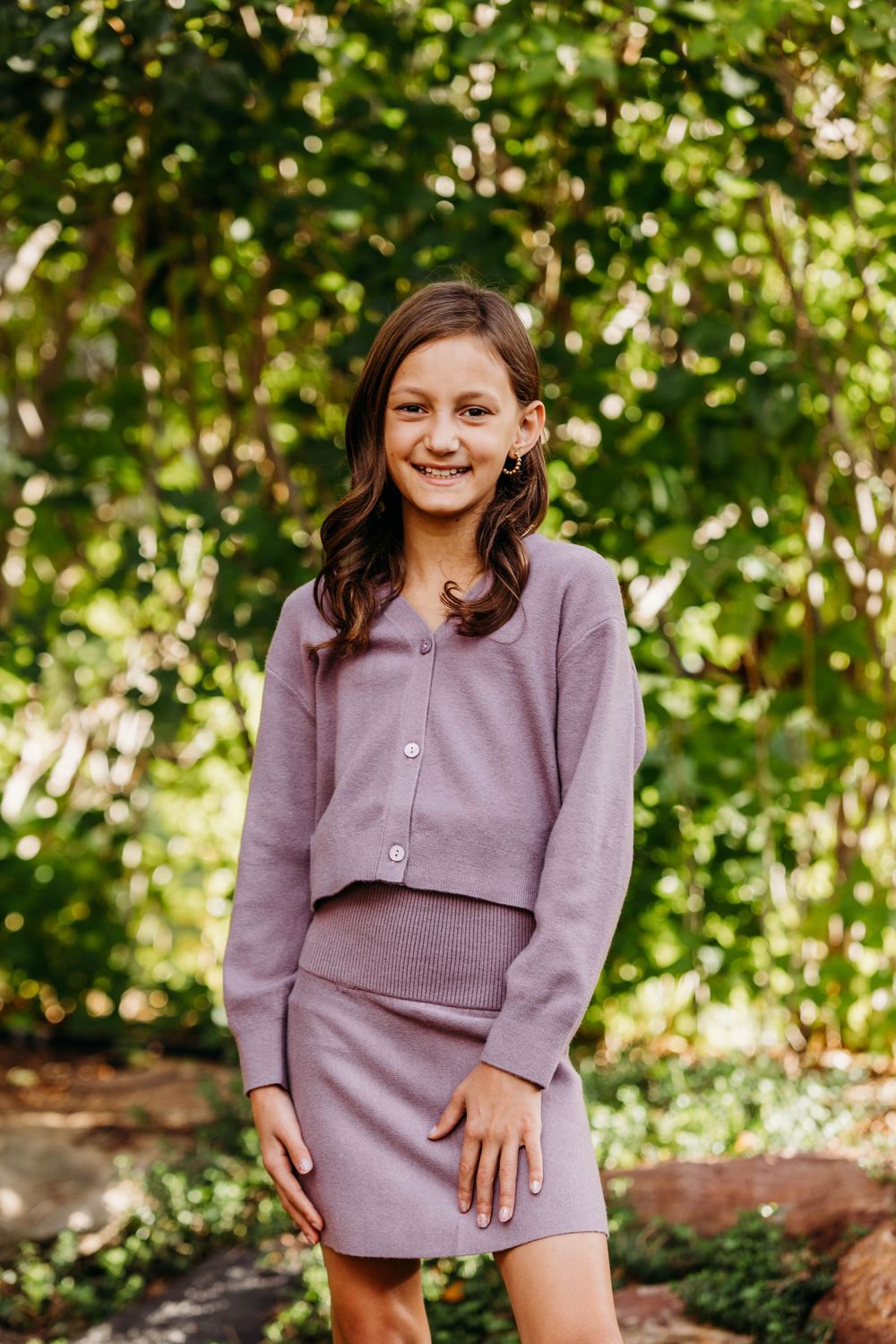 Young girl in lavender knit skirt suit smiling outdoors with green foliage background.