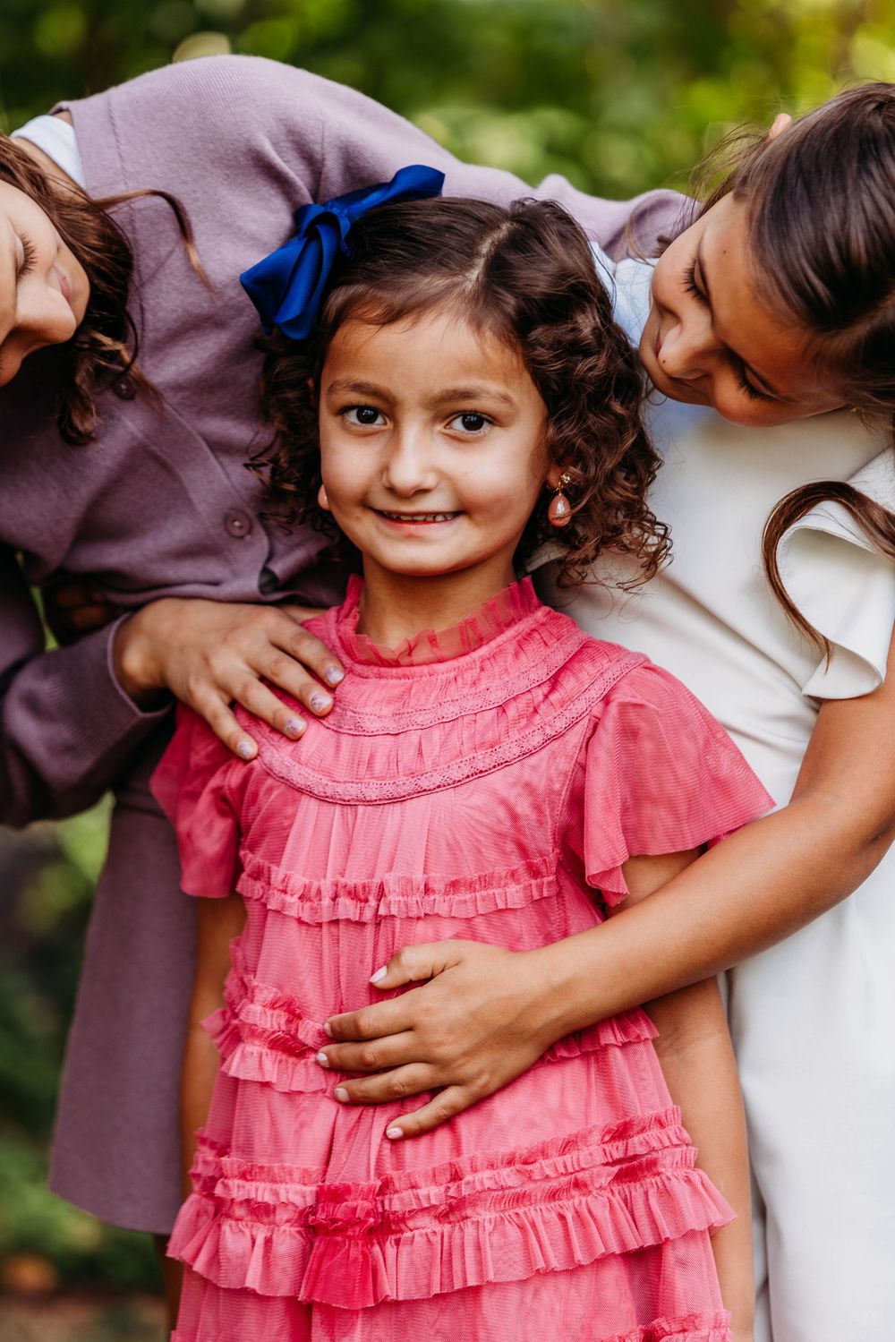 Young girl in pink ruffled dress smiles with blue bow, flanked by two siblings hugging her outdoors.