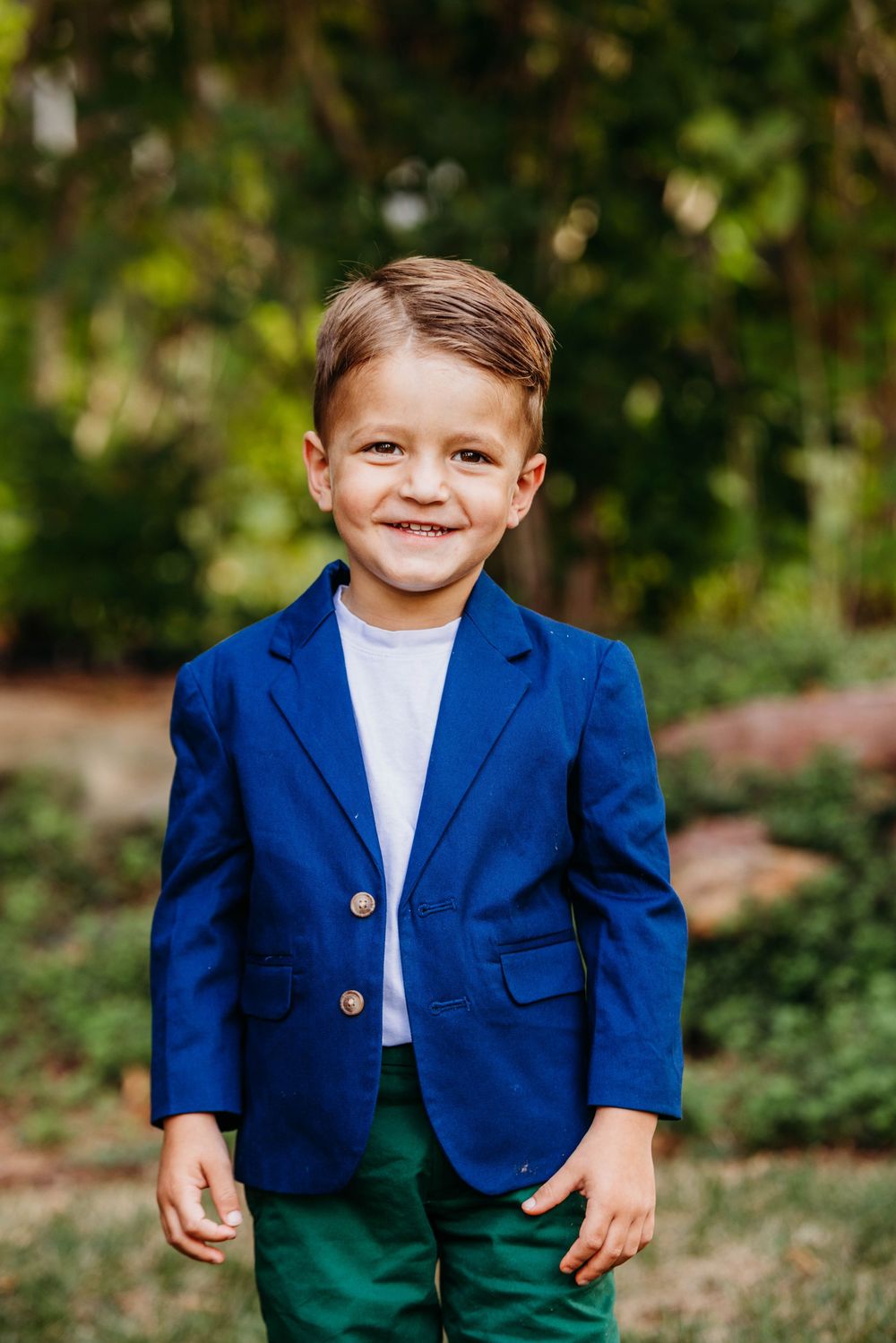 Smiling young boy in a blue blazer and green pants poses outdoors in a lush green garden setting.
