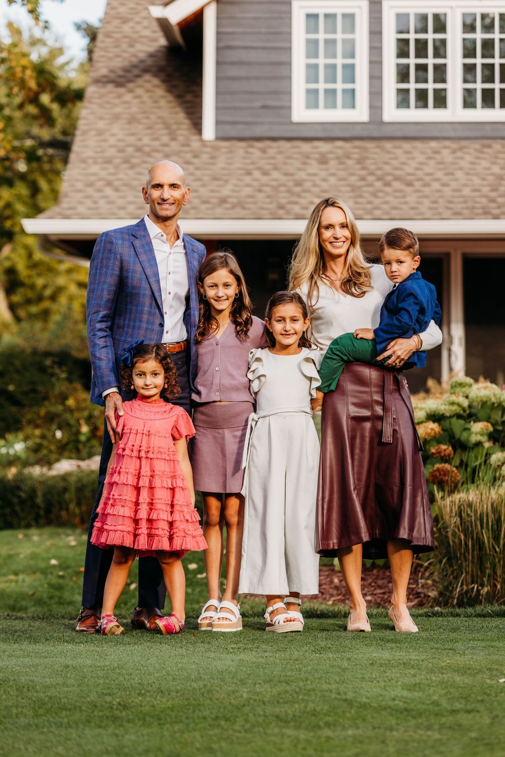 Happy family of six posing outdoors in front of a large home, dressed in elegant fall attire on a green lawn.
