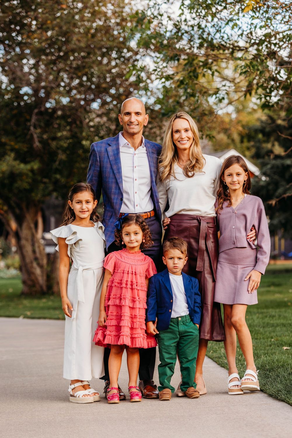 Family of six posing outdoors, parents with four children dressed in elegant attire, smiling in a lush green park setting.