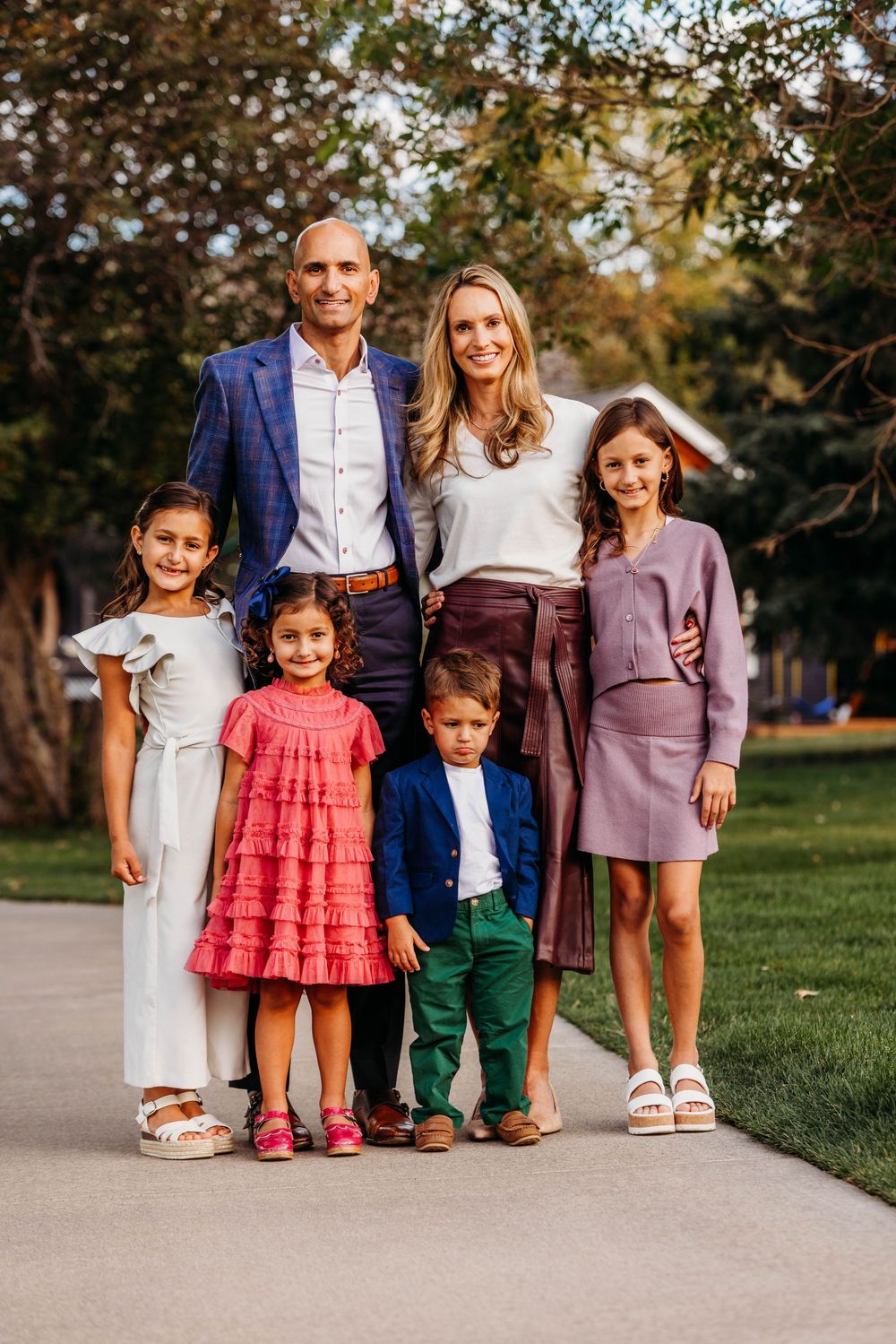 A happy family of six poses outdoors on a sunny day, dressed in elegant semi-formal attire on a tree-lined path.