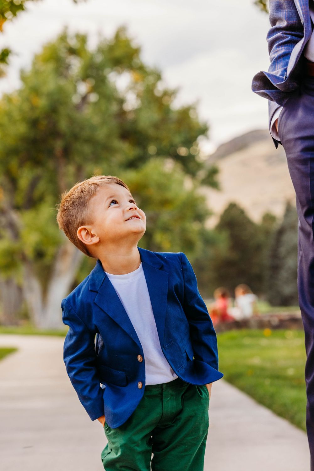 A young boy in a blue blazer and green pants looks up adoringly at an adult in a sunny park setting.