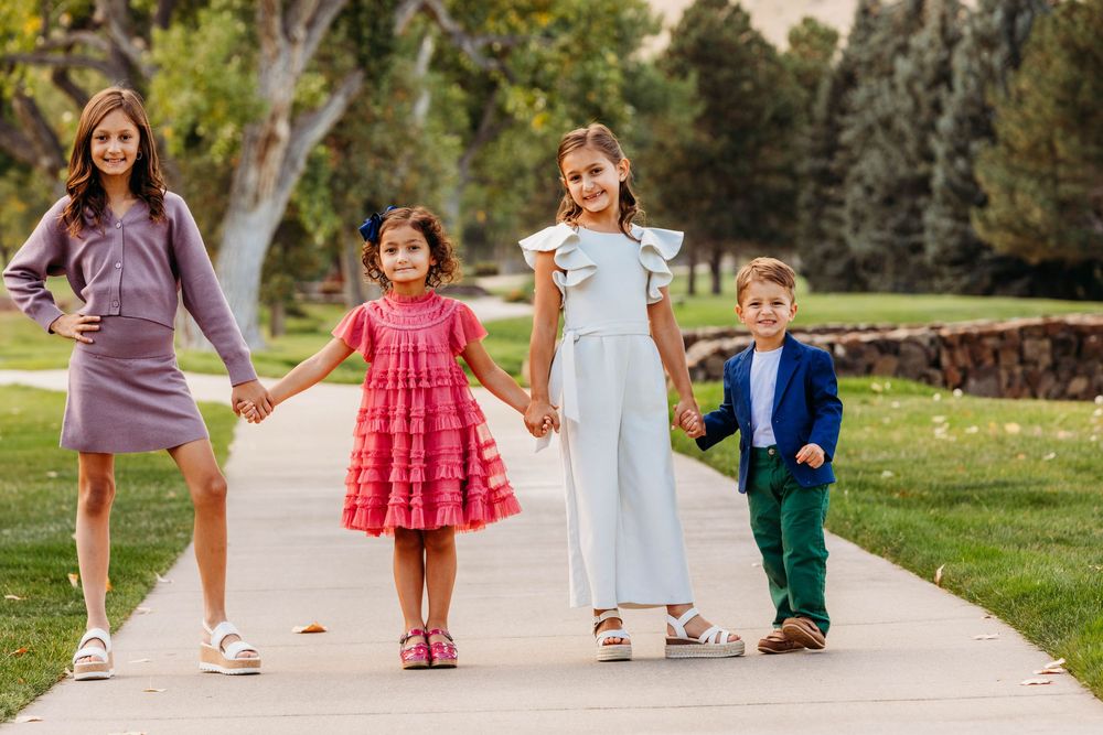 Four children in dressy outfits hold hands on a park path, smiling for a family portrait outdoors.
