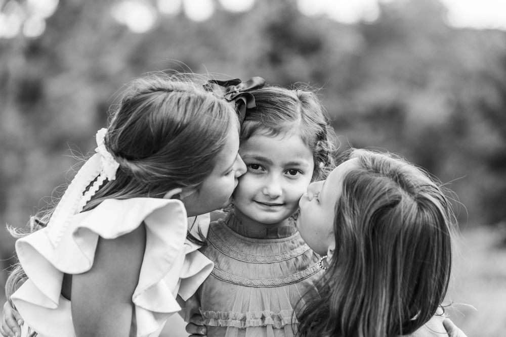 Two girls kissing a smiling young girl on the cheeks in a sweet outdoor black and white portrait photo.