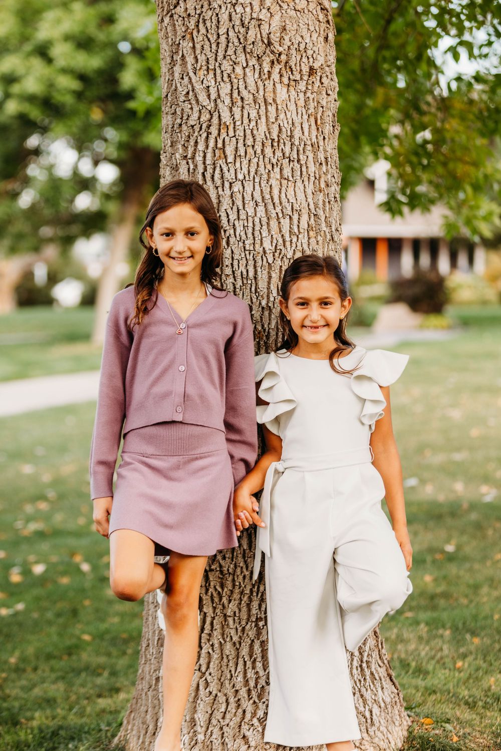 Two smiling girls in stylish outfits leaning against a tree in a park.