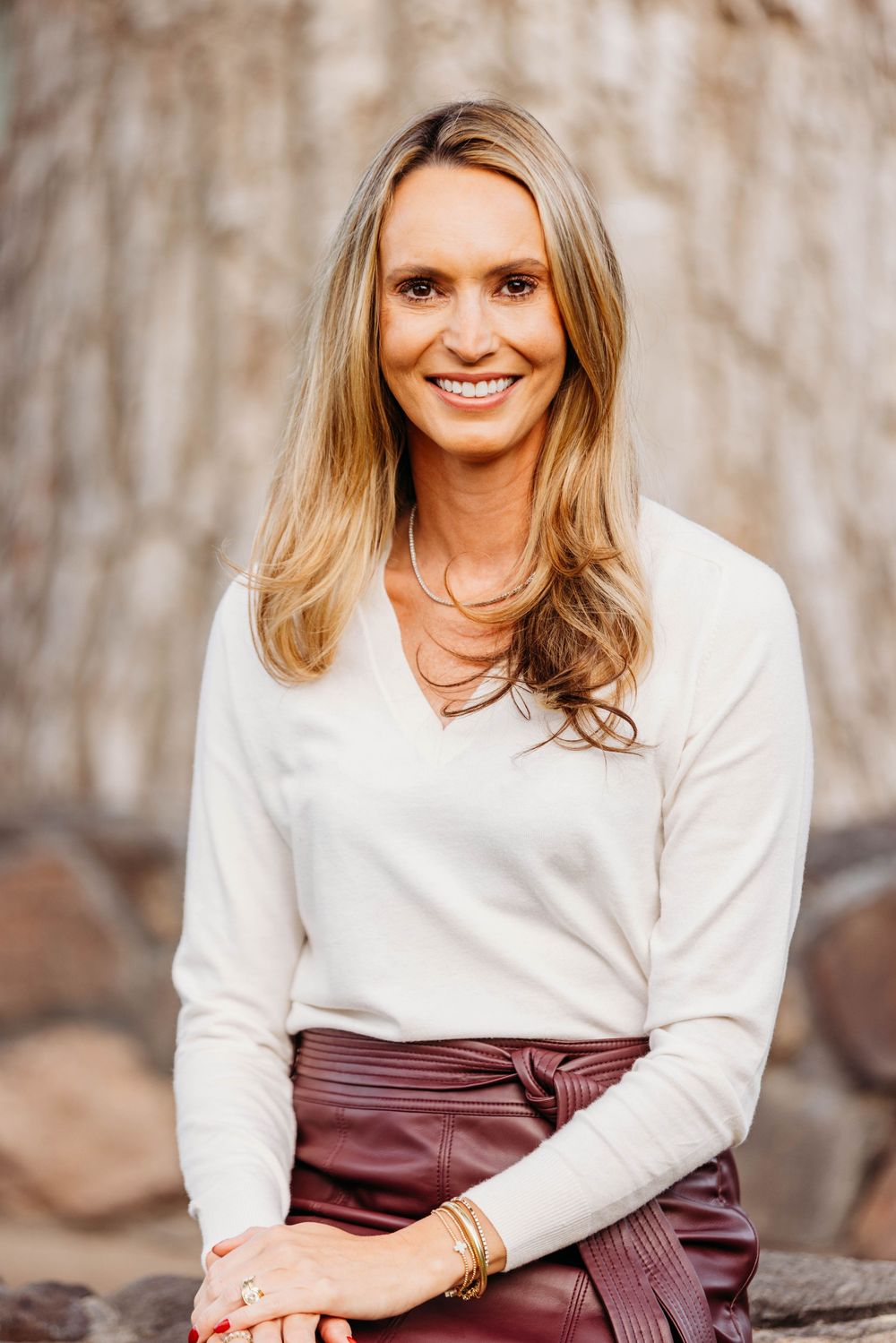 Smiling blonde woman in white sweater and burgundy leather skirt seated outdoors against stone wall.
