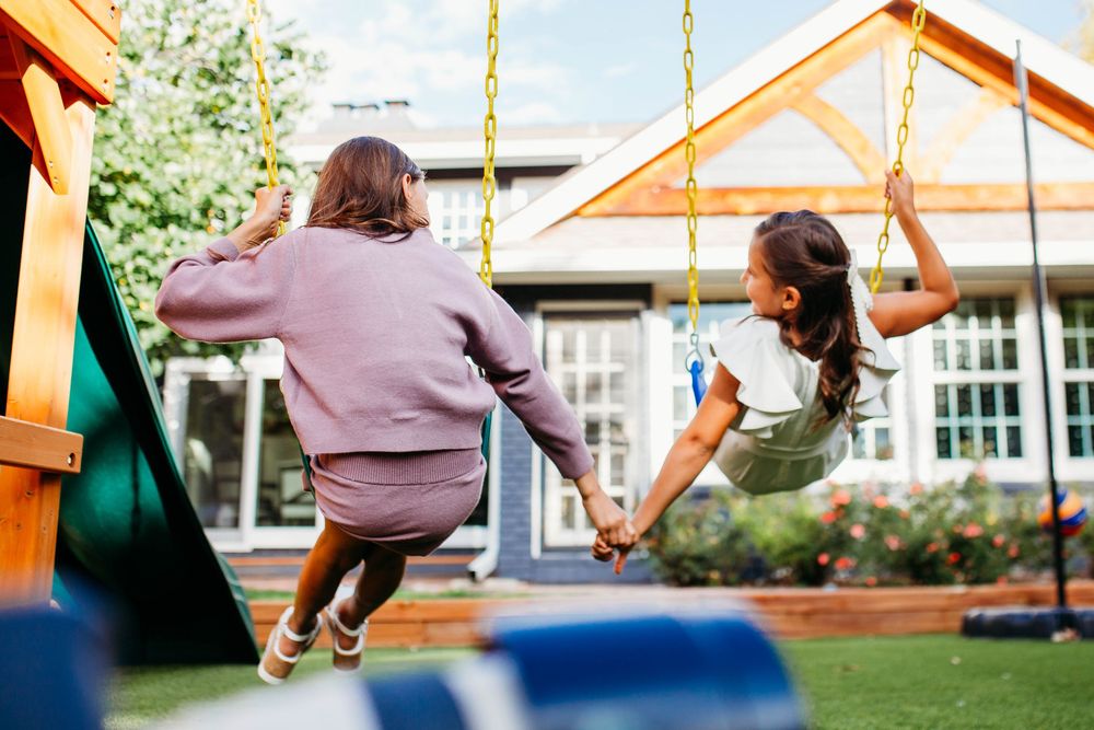 Two girls holding hands while swinging on backyard playground swings, enjoying a sunny day together.