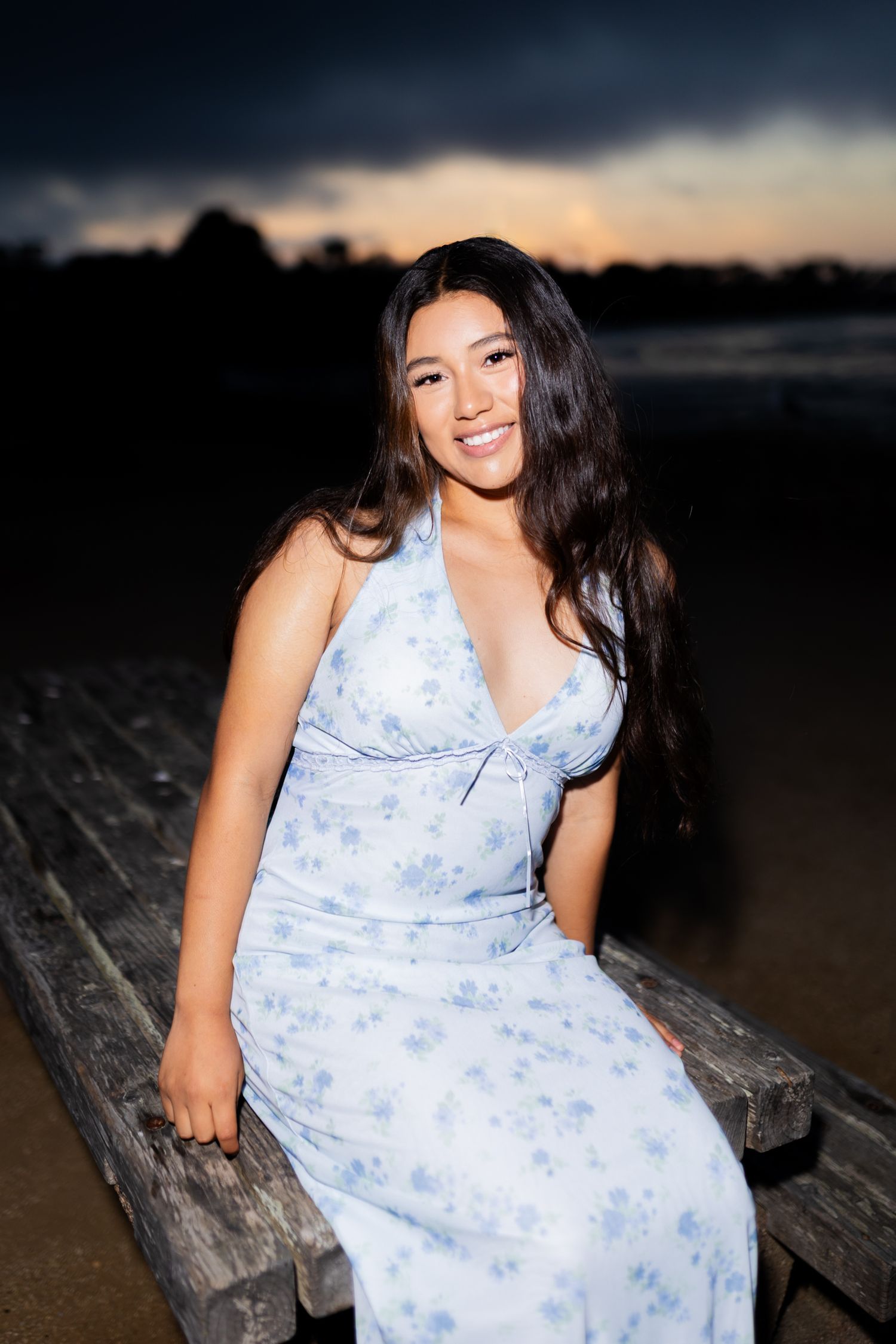 A smiling young woman in a floral blue dress sits on a wooden dock at sunset with a dramatic sky behind her.