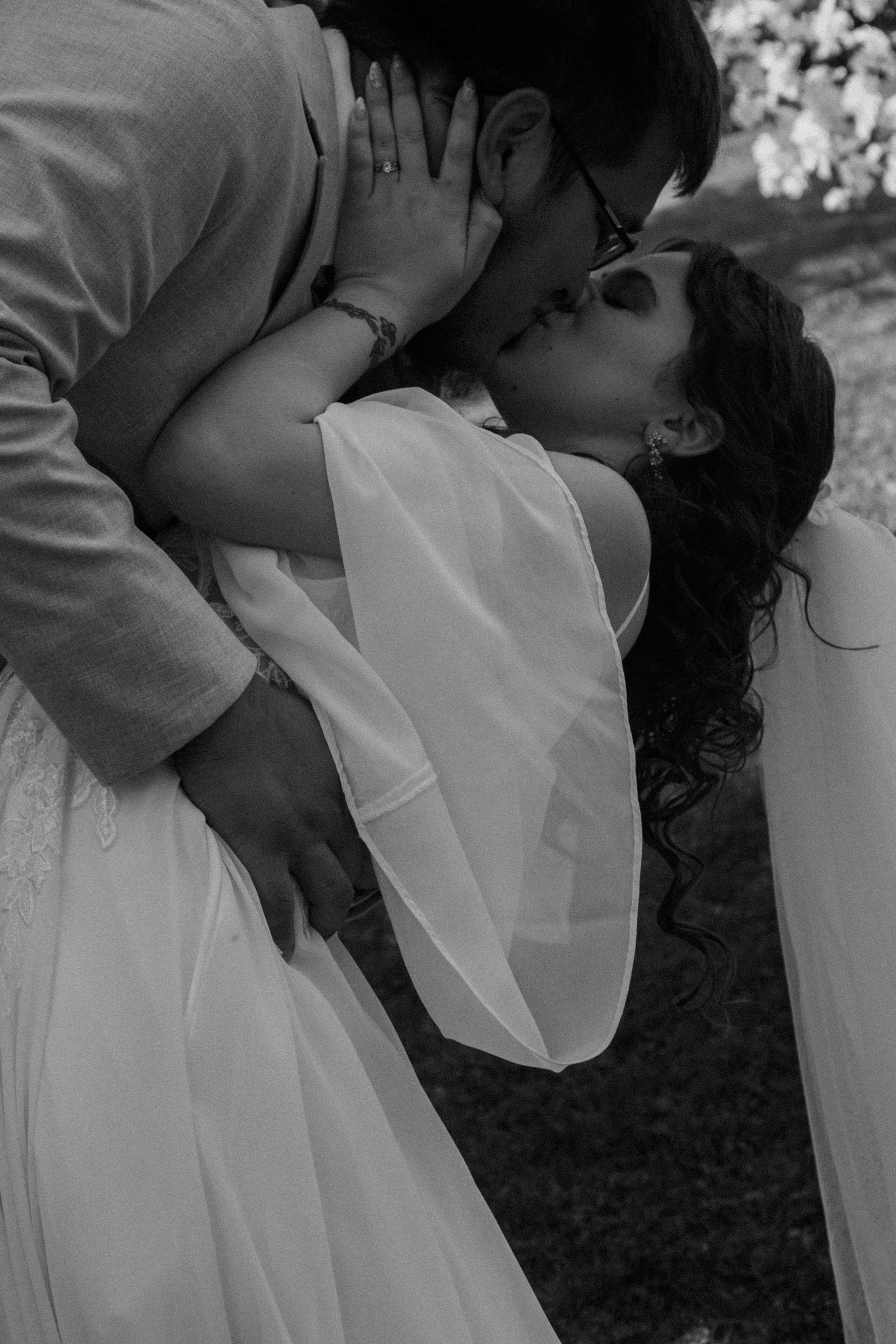 Black and white photo of a bride and groom embracing outdoors, her lace gown flowing in the wind.