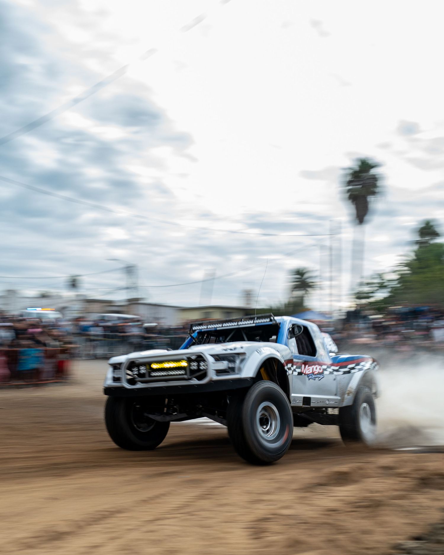 A white off-road trophy truck races on a dirt track at a desert rally event with crowds watching