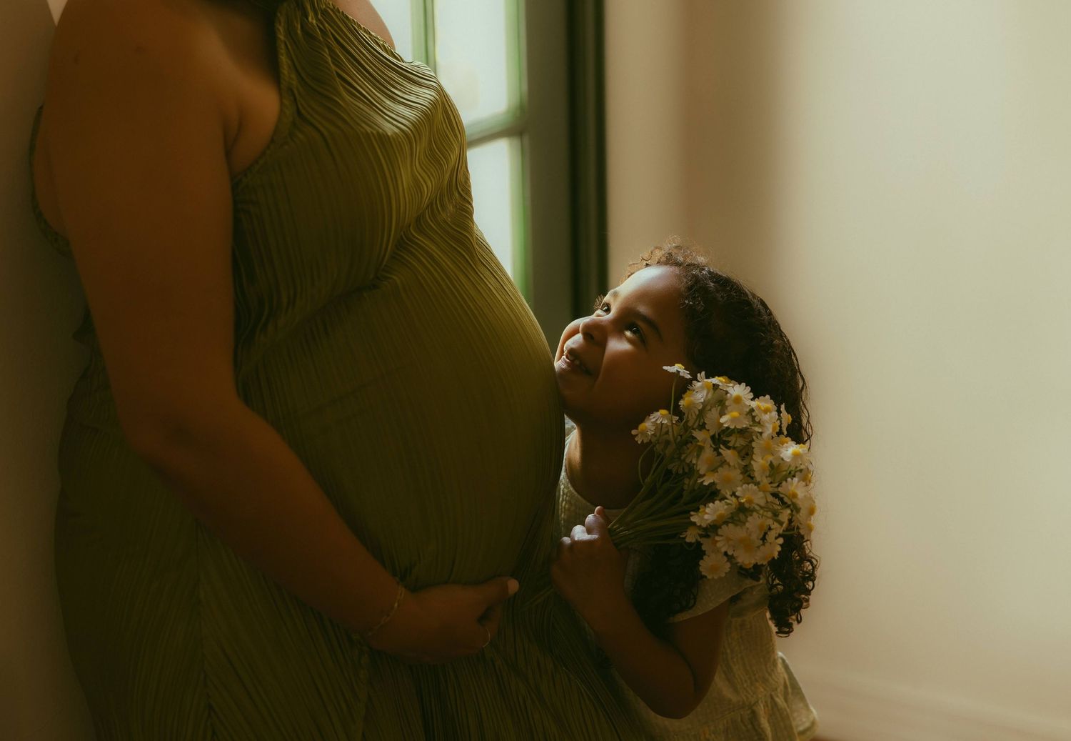 A young girl holding white flowers looks up at her pregnant mom in a green dress standing by a window. Family photo poses orange county