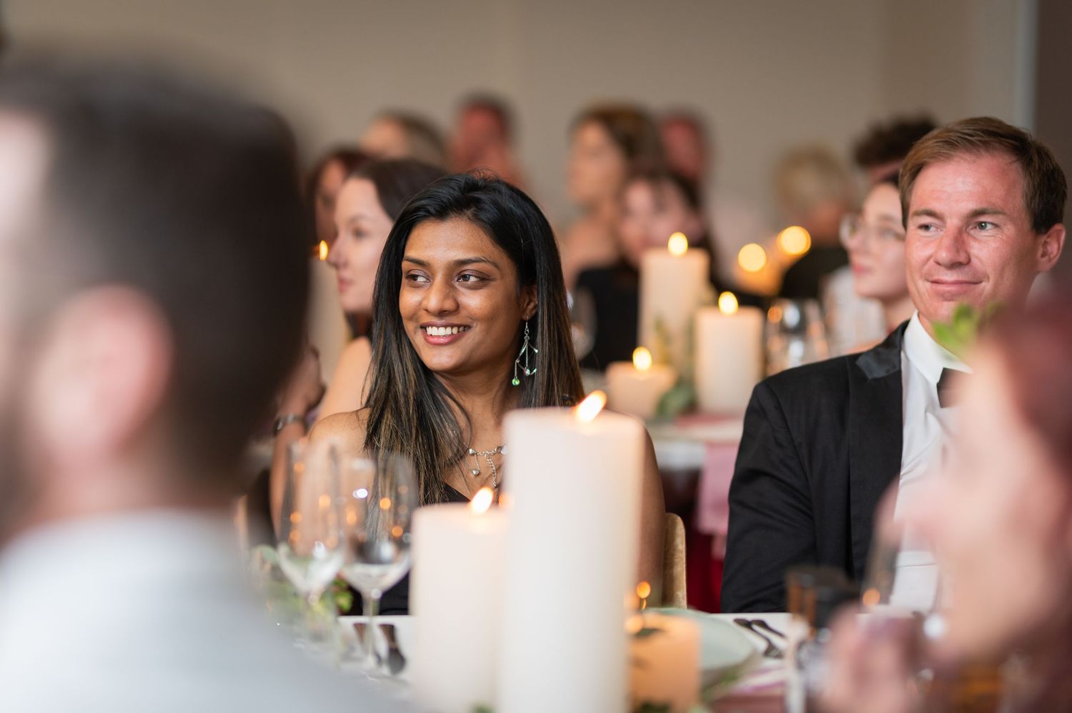 Guests smiling and enjoying a candlelit formal dinner event, with candles and wine glasses on the table.
