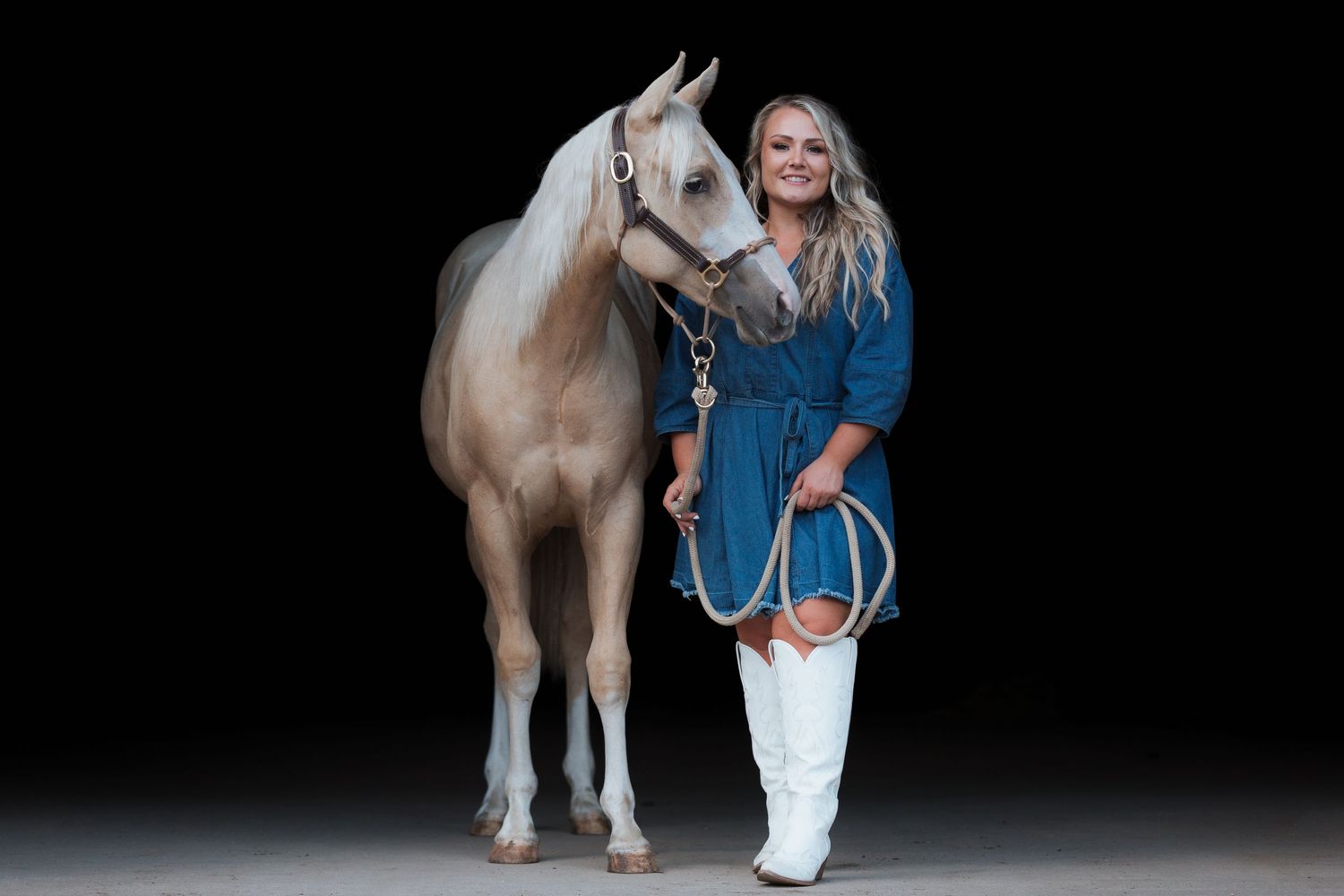 Woman in blue dress and white boots poses with a white horse against a dark studio background.