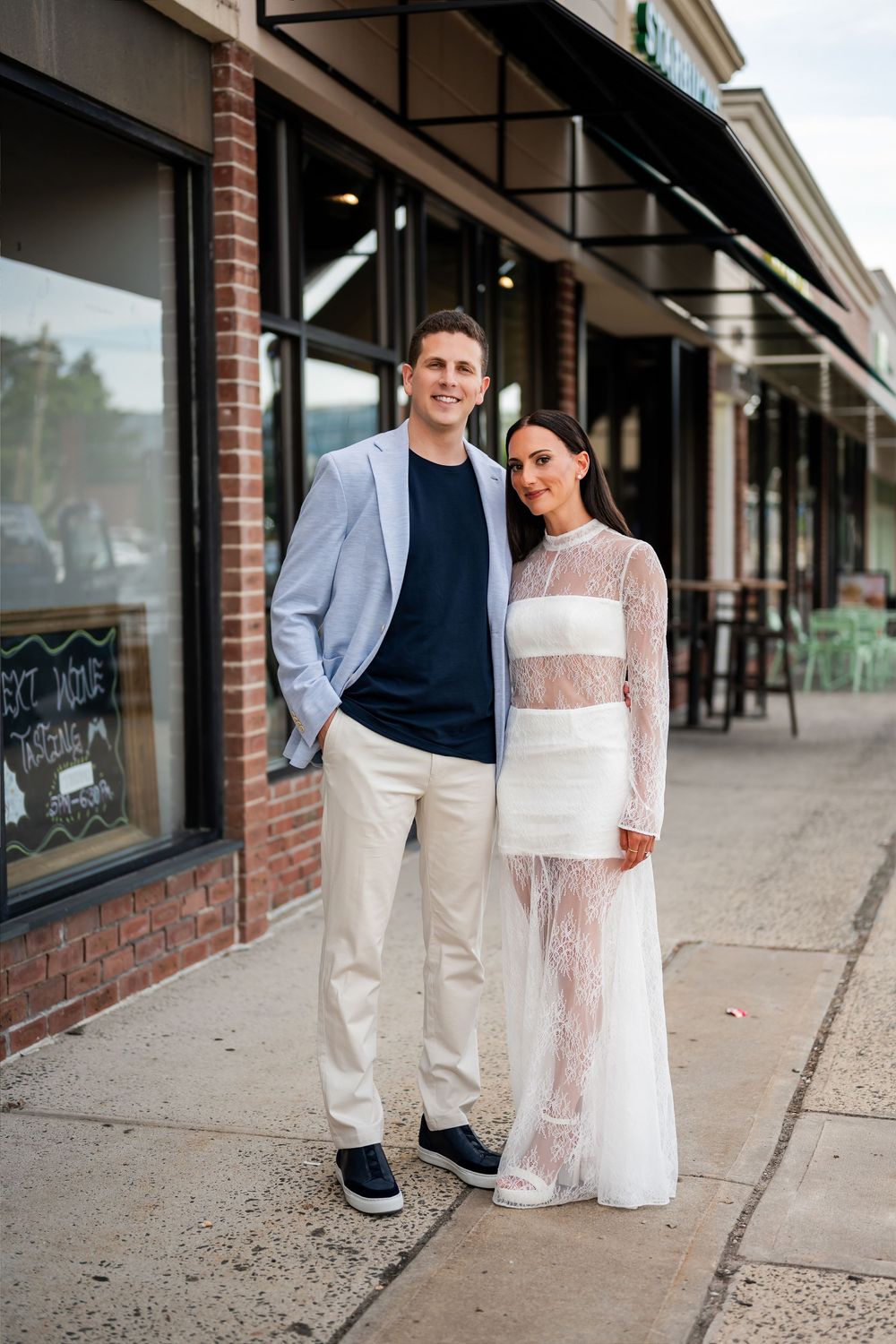 Couple posing on sidewalk outside brick storefront, man in blazer and chinos, woman in white lace gown.