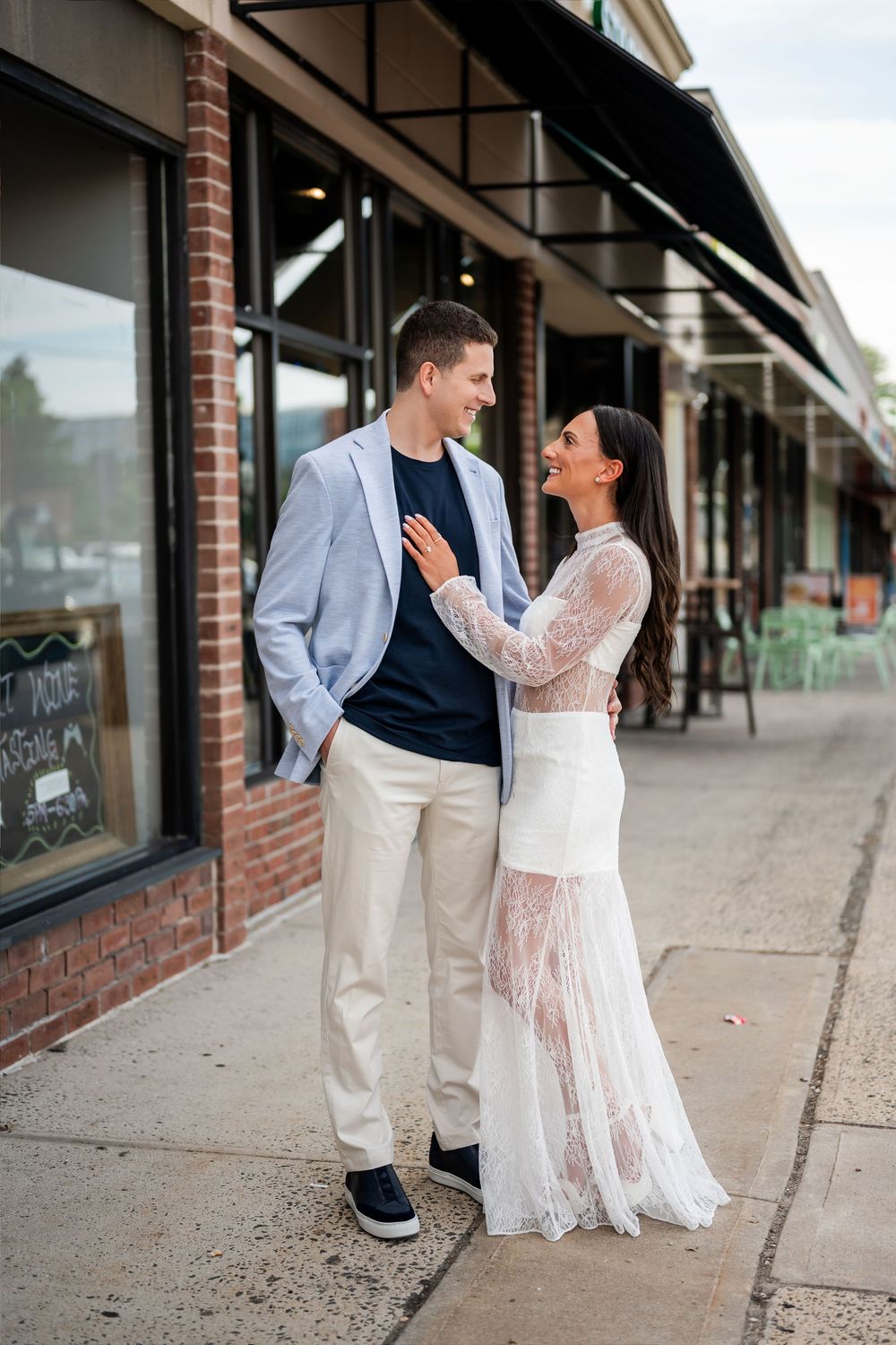 A couple in elegant attire smiling at each other on a brick sidewalk outside a charming storefront.