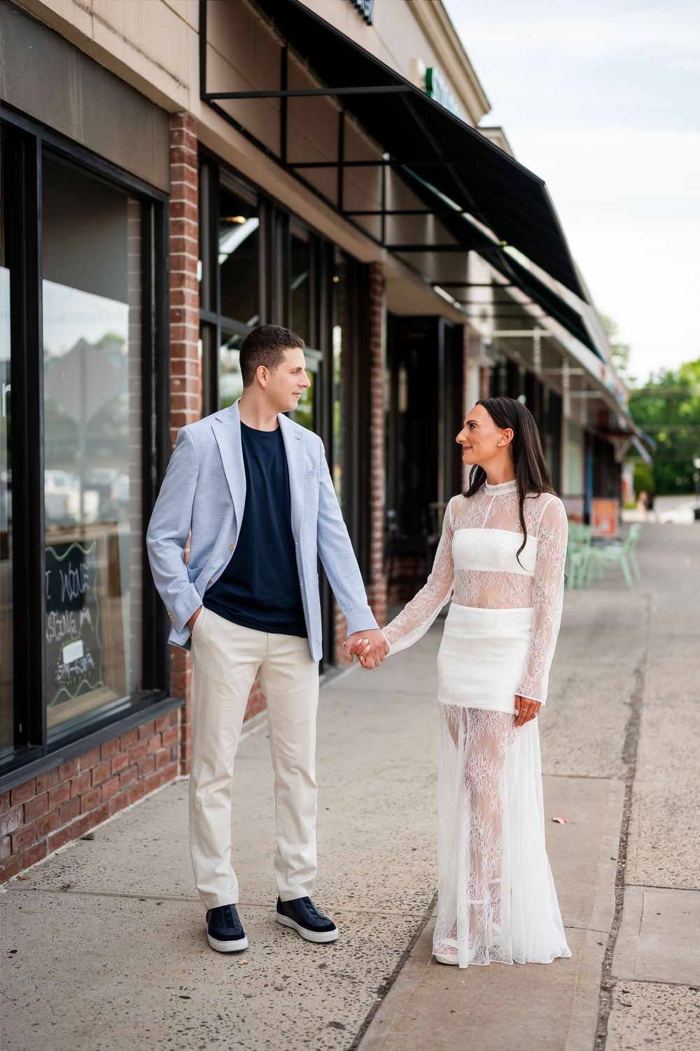 Couple holding hands in front of brick storefront, man in blazer and woman in white lace dress.