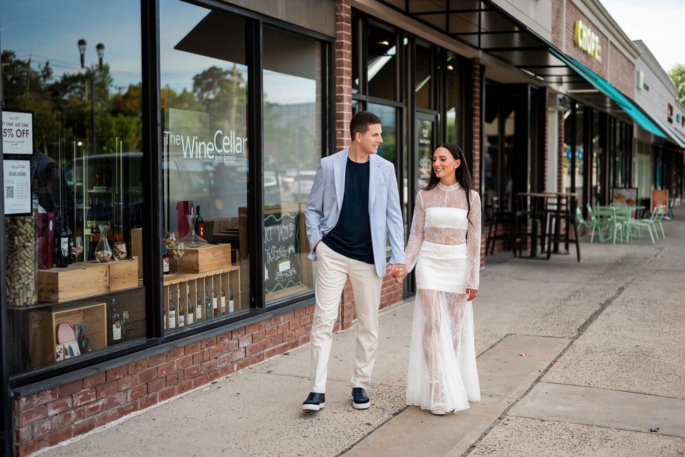 Couple holding hands walking past a wine cellar storefront on a sunny day, man in blazer and woman in white lace dress.