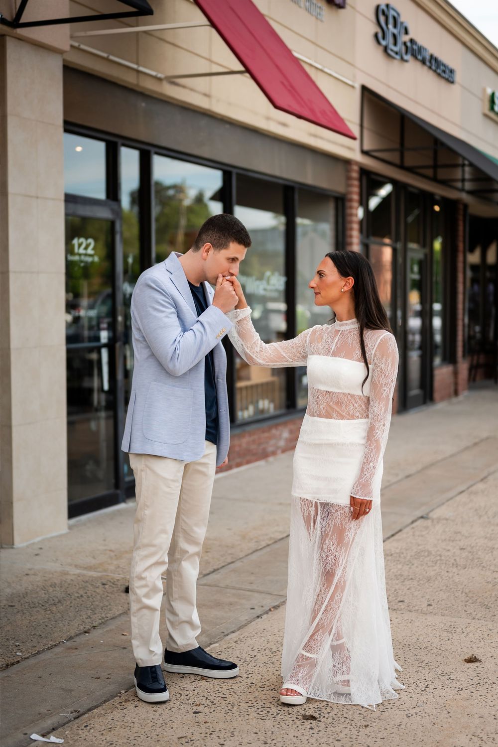 A man in a blue blazer kisses a woman's hand as she wears a white lace dress outside a shopping center.