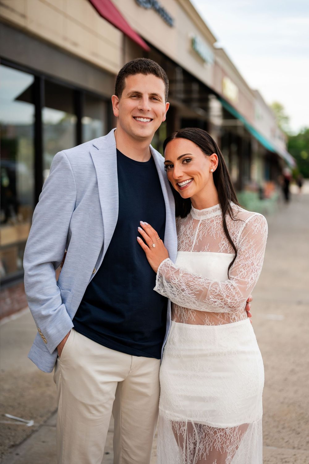 Smiling couple posing outdoors, man in light blazer and navy shirt, woman in white lace dress, near storefronts.