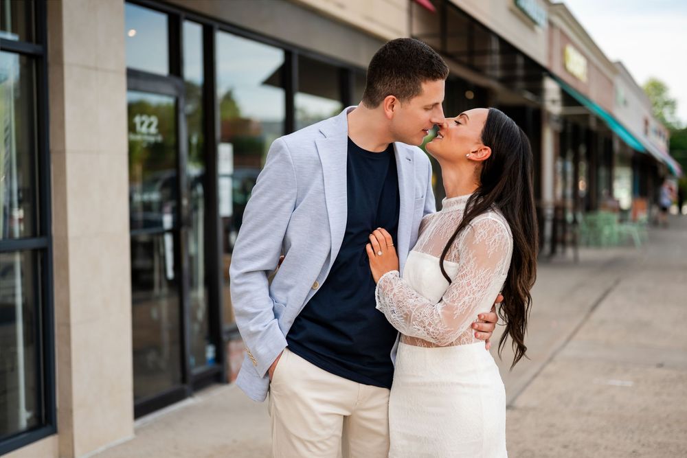 Couple sharing a romantic kiss outside a shopping center, man in blazer and woman in white lace dress.