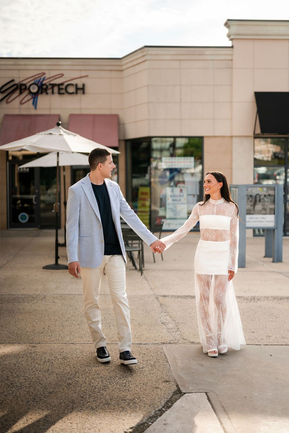 Couple holding hands walking outside a shopping center, woman in white lace dress, man in light blazer and khakis.