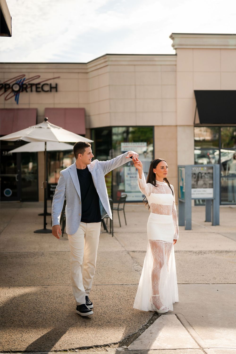A couple dances elegantly outside a shopping center, man in gray blazer twirling woman in white sheer dress.