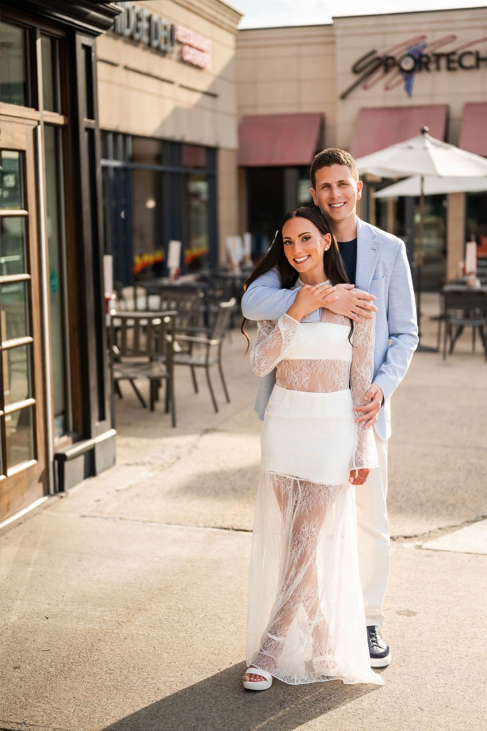 Couple posing outside shops, woman in white lace dress, man in light blue blazer, smiling together on sidewalk.