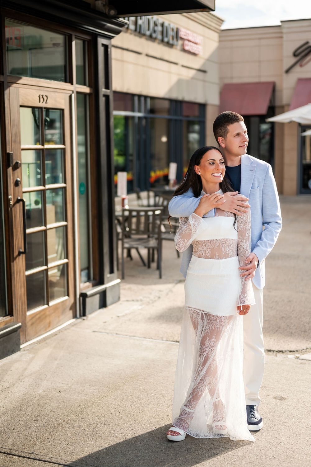 Couple posing outside a restaurant, woman in white lace dress, man in light blue suit, urban setting.