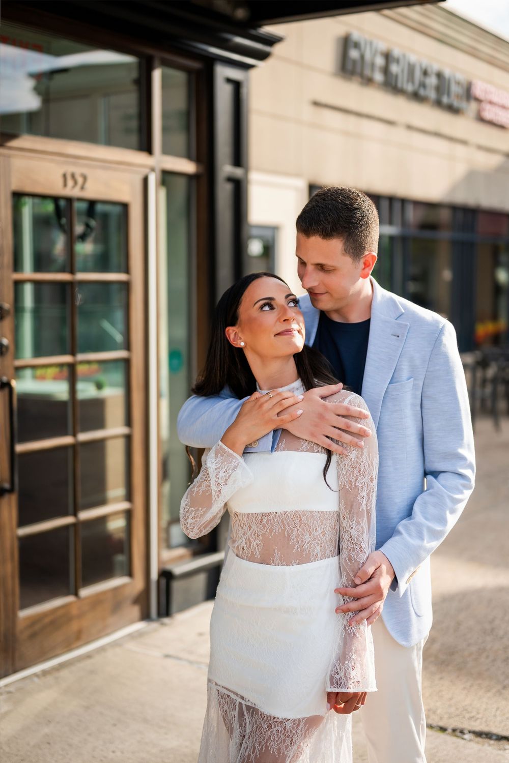 Couple embracing outside a restaurant, woman in white lace dress gazing up at man in light blue blazer.