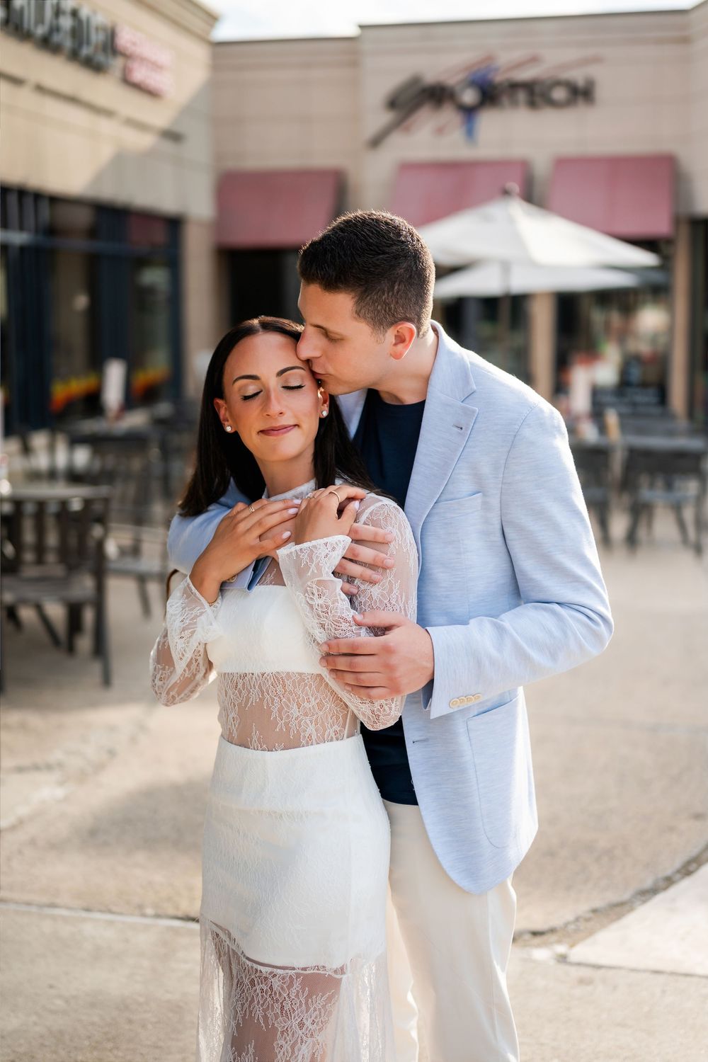 A couple shares a tender moment outdoors, man in light blazer kisses woman in white lace dress near a shopping center.