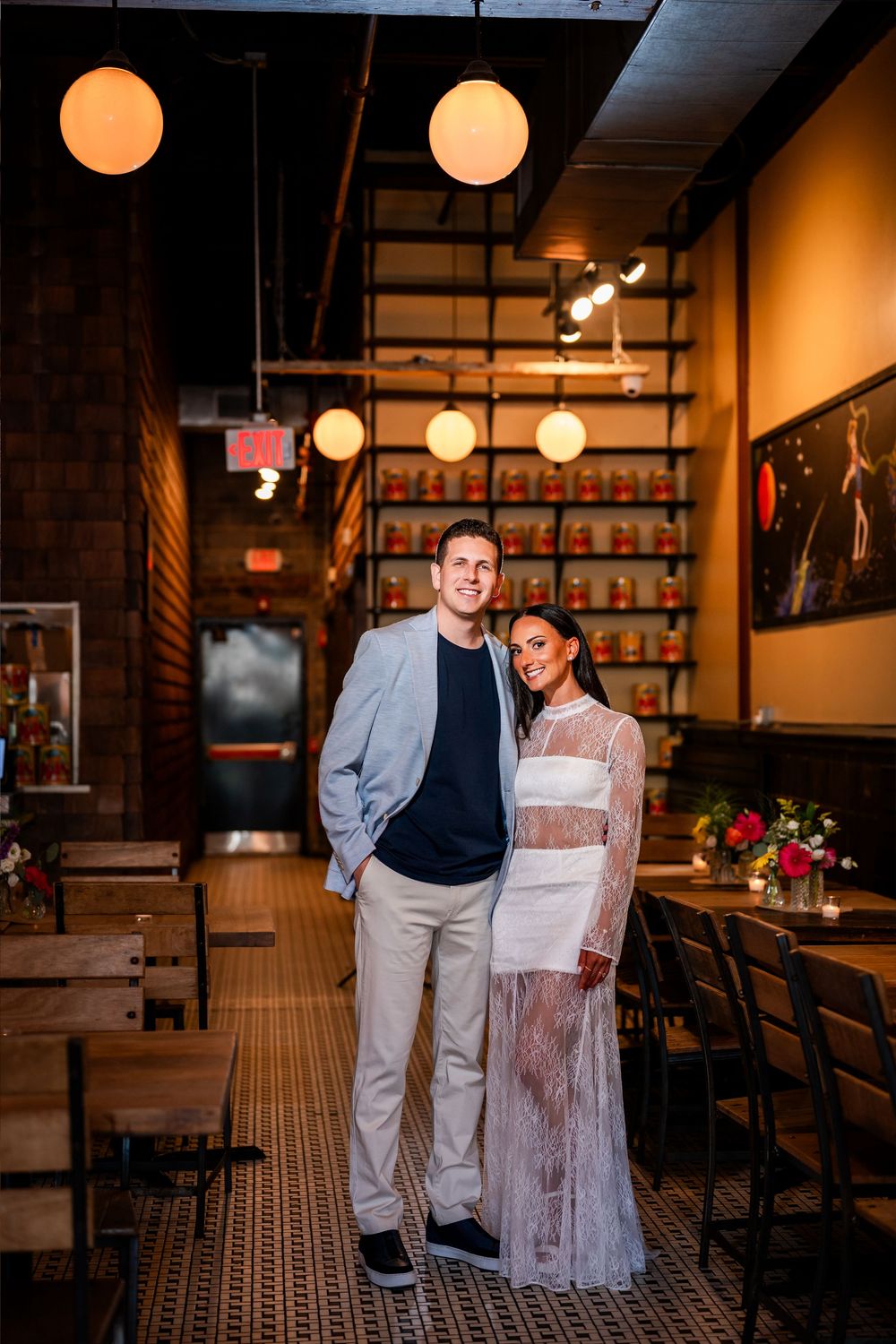 Couple posing together in a warmly lit restaurant interior with globe pendant lights and wooden chairs.