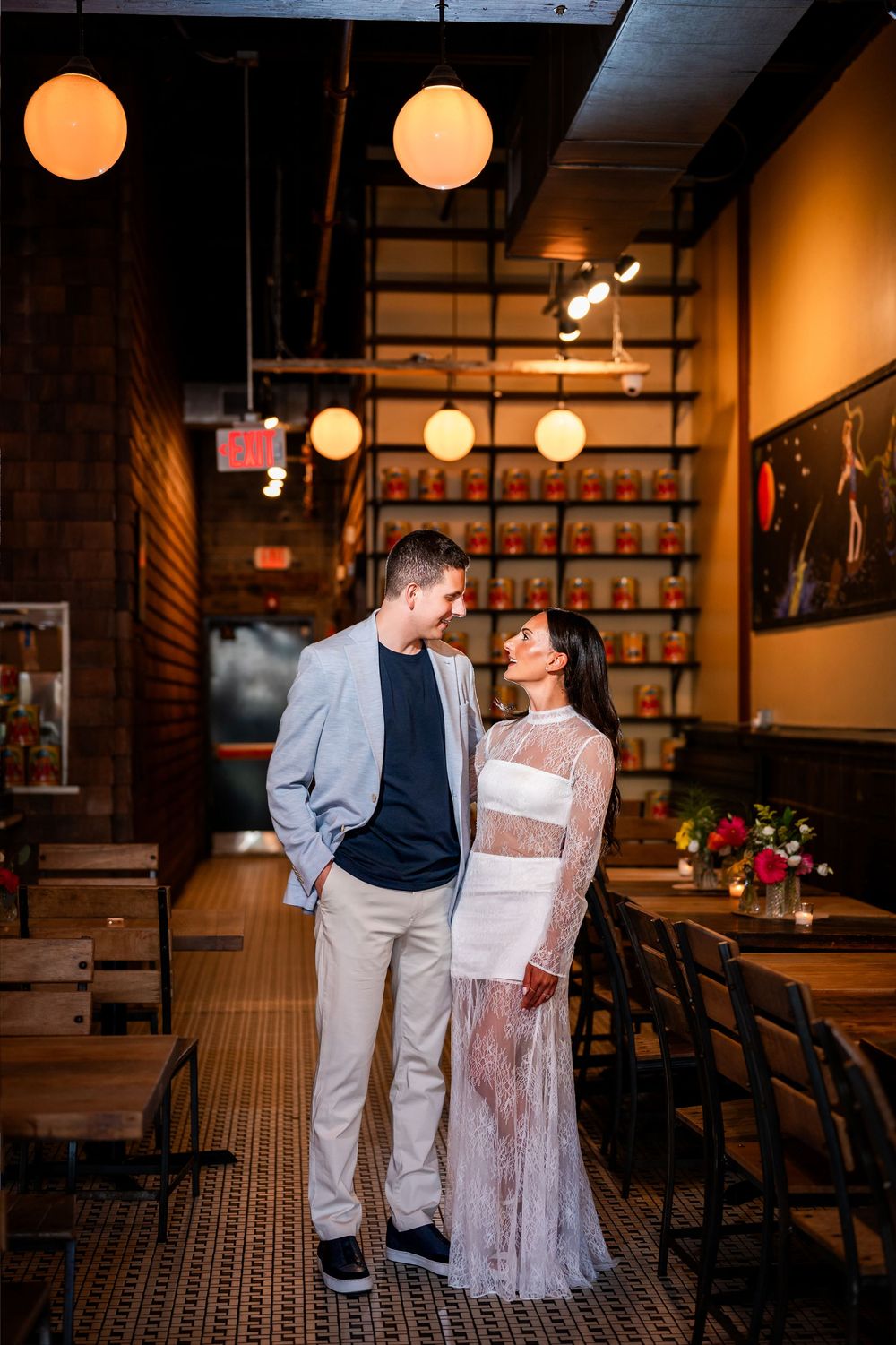 Couple sharing a romantic moment in a warmly lit restaurant, woman in lace dress, man in gray blazer.