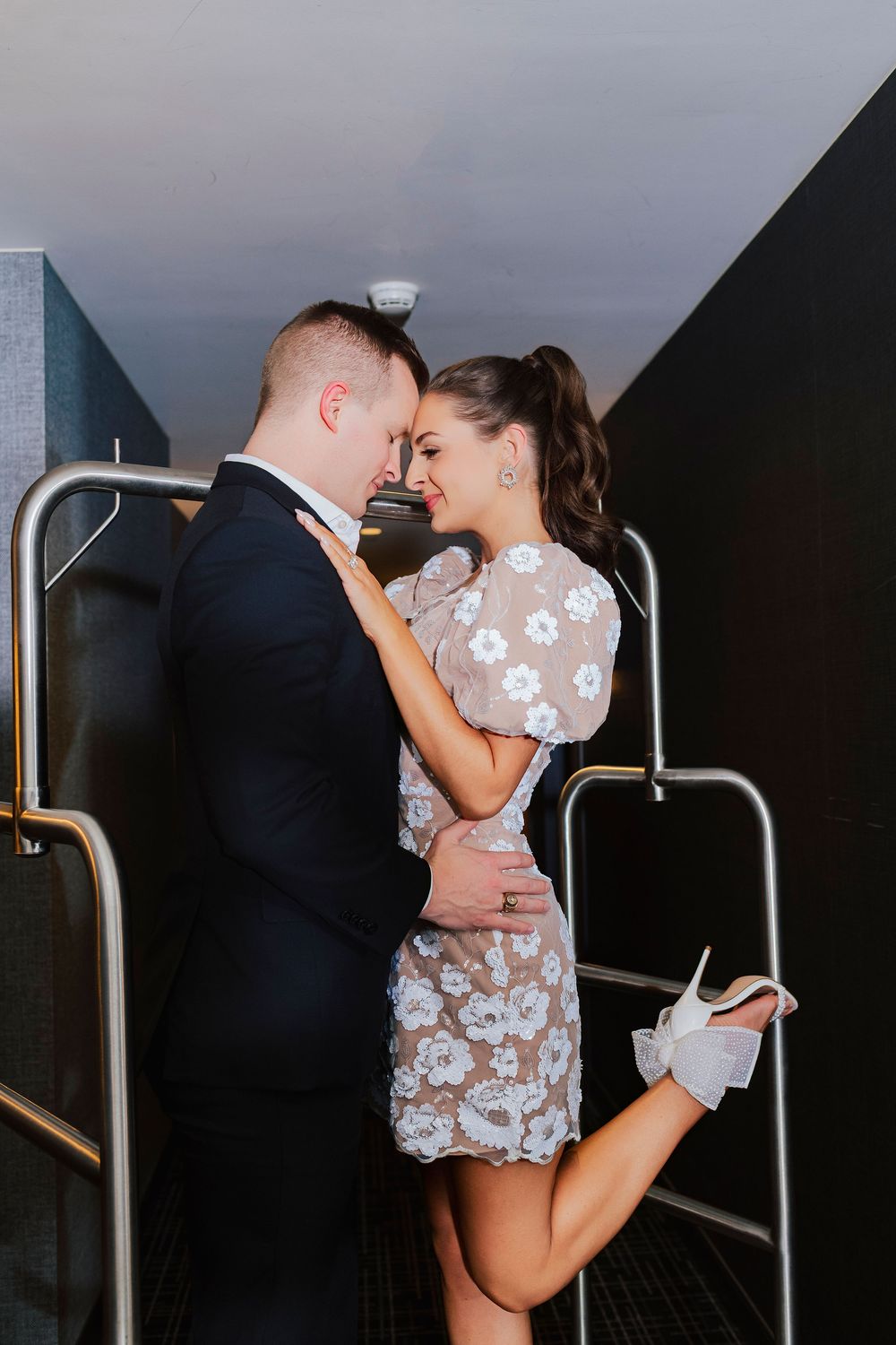 Couple in formal attire sharing a romantic moment on stairs, woman in floral mini dress, man in black suit.
